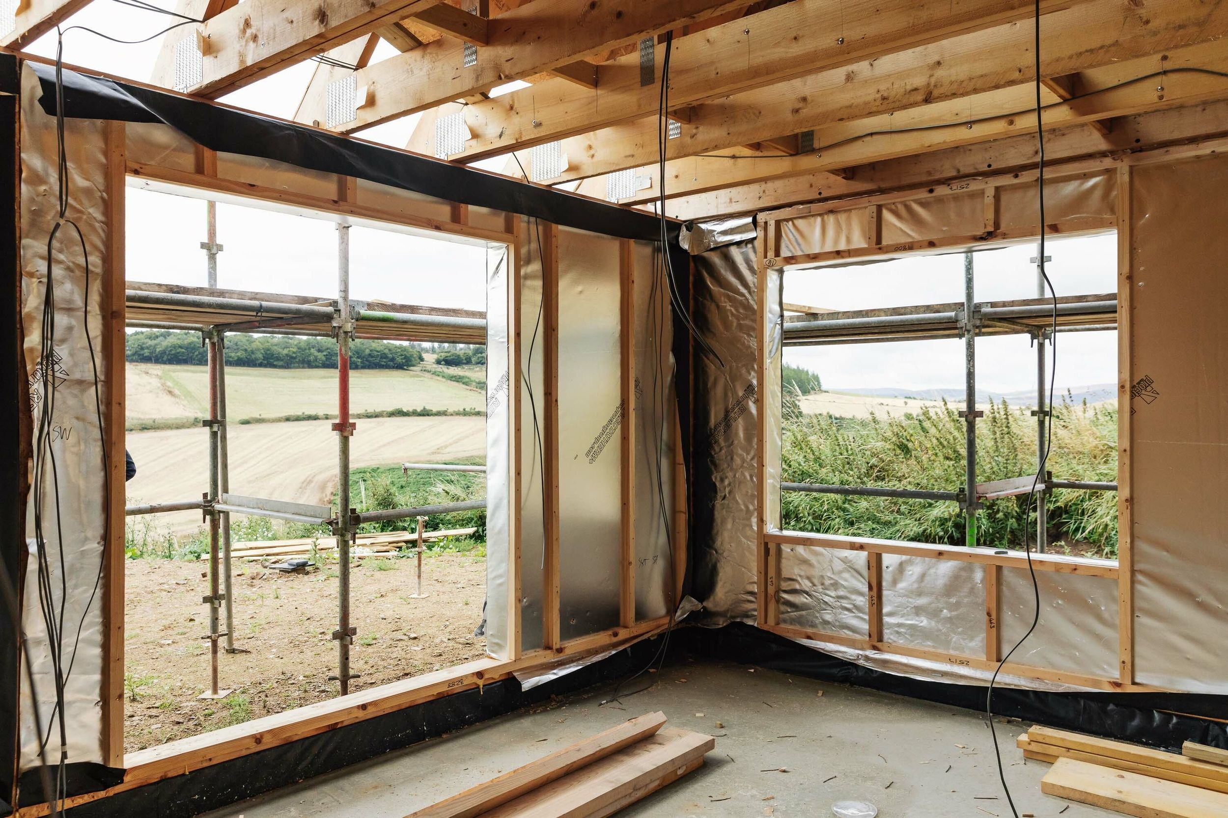 Inside a half-built timber frame home. Materials include Oriented Strand Board (OSB). One wall of the new house is covered in silver, windproof building paper.  Timber frames are exposed. Electrical cables hang from the ceiling. Sawdust covers the concrete floor. Barley fields are in the distance.