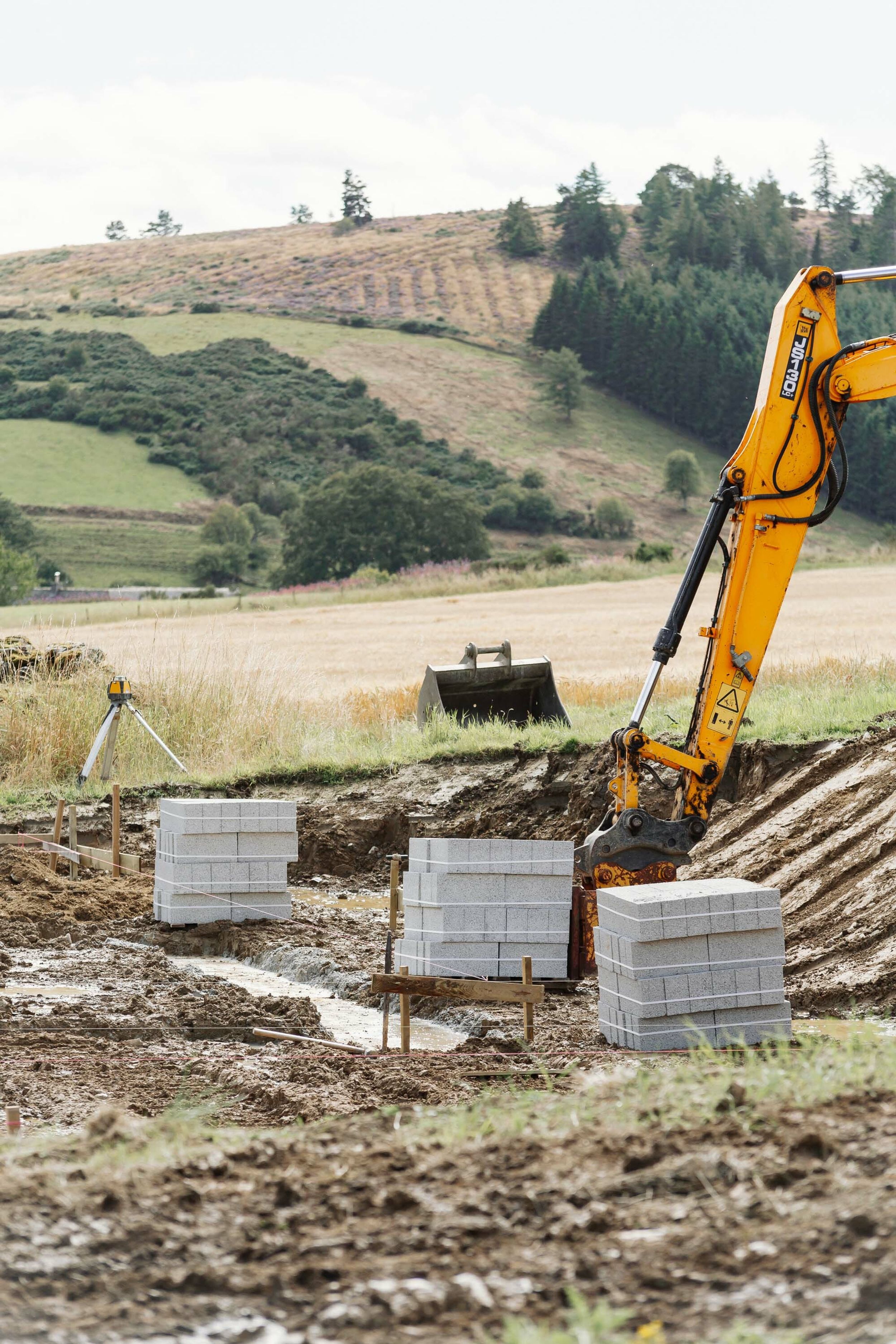 Stacks of grey concrete blocks sit next to the muddy foundations of new home. Builders line has been run around the permitter of the foundations. A yellow digger arm is visible. A digger bucket and laser level are in the background. A forest covered hill is in the distance.