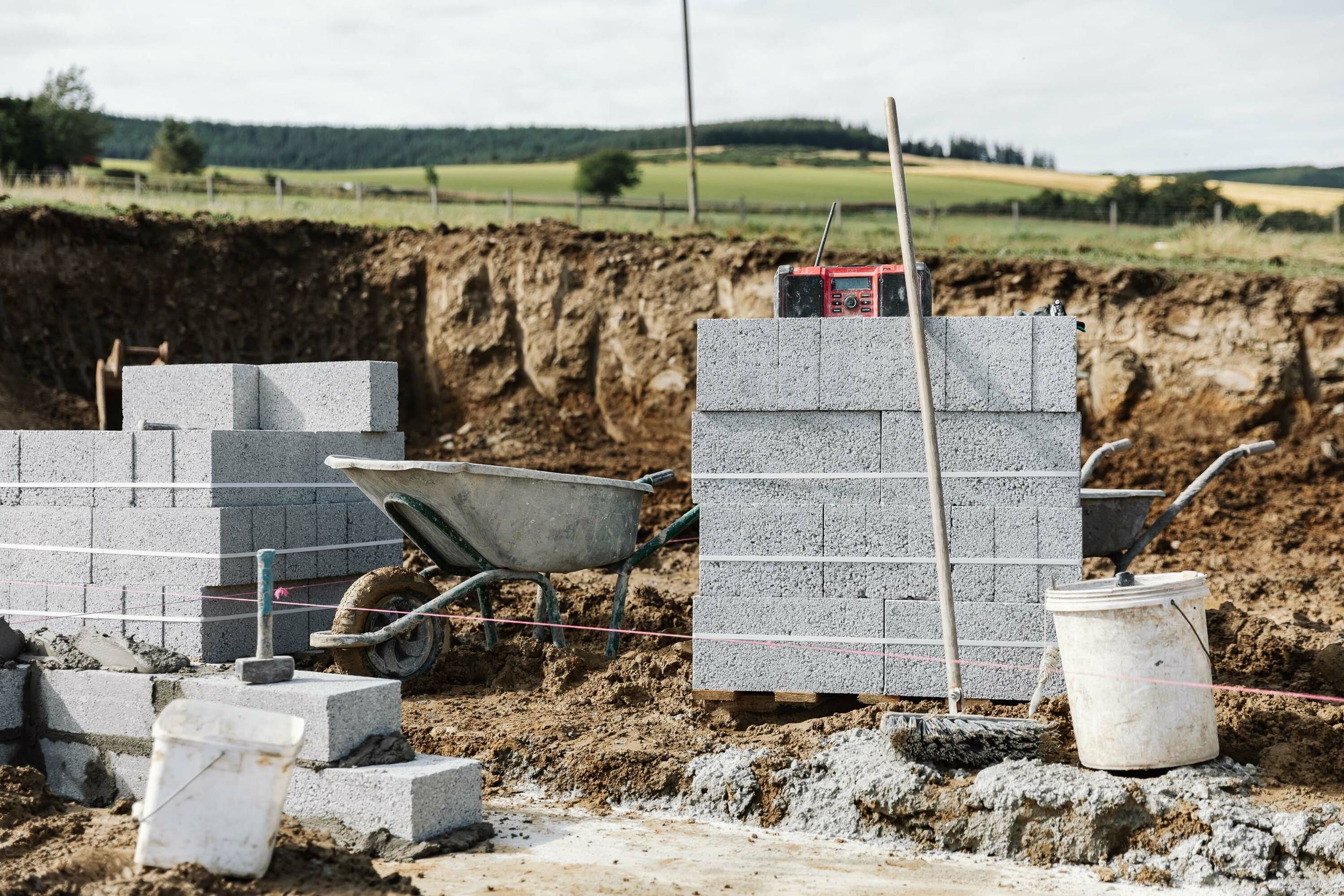 A large pallet of concrete builder’s blocks sits on a muddy construction site. A red radio sits atop of the concrete blocks. A cement splattered barrow and a white bucket sit close by. A broom leans against the blocks. Barley fields can be seen in the distance.