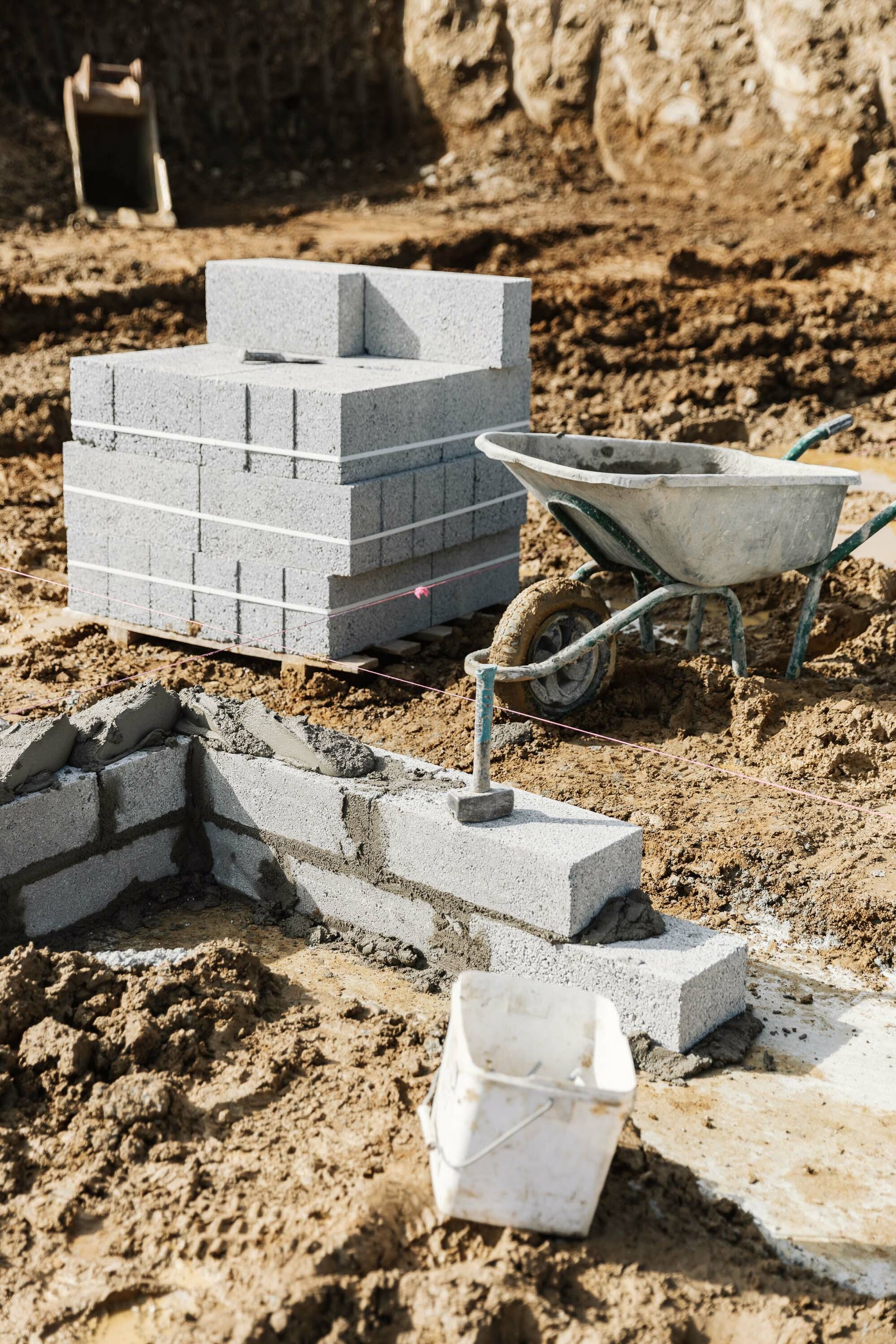 A pallet of builder’s blocks sits on a muddy construction site. A wheelbarrow sits next to the blocks. In the foreground, a new wall is being built from concrete blocks on the foundations of a new house.