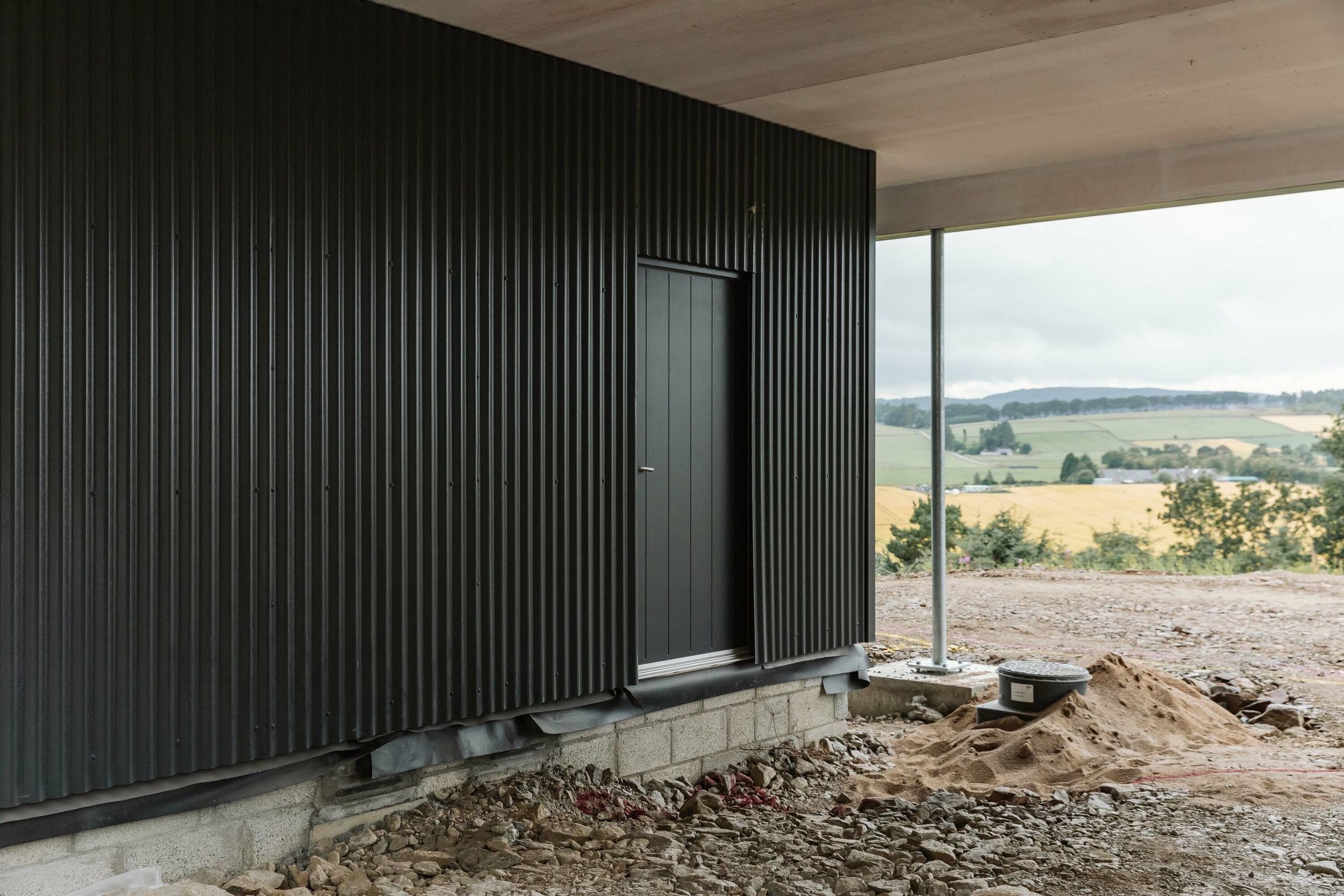 A partially-built home clad in grey corrugated sheeting. A black door is in the middle of the sheeting. Beyond the home, there is rural views of farmer’s fields. Rubble and builder’s sand can be seen in the foreground.