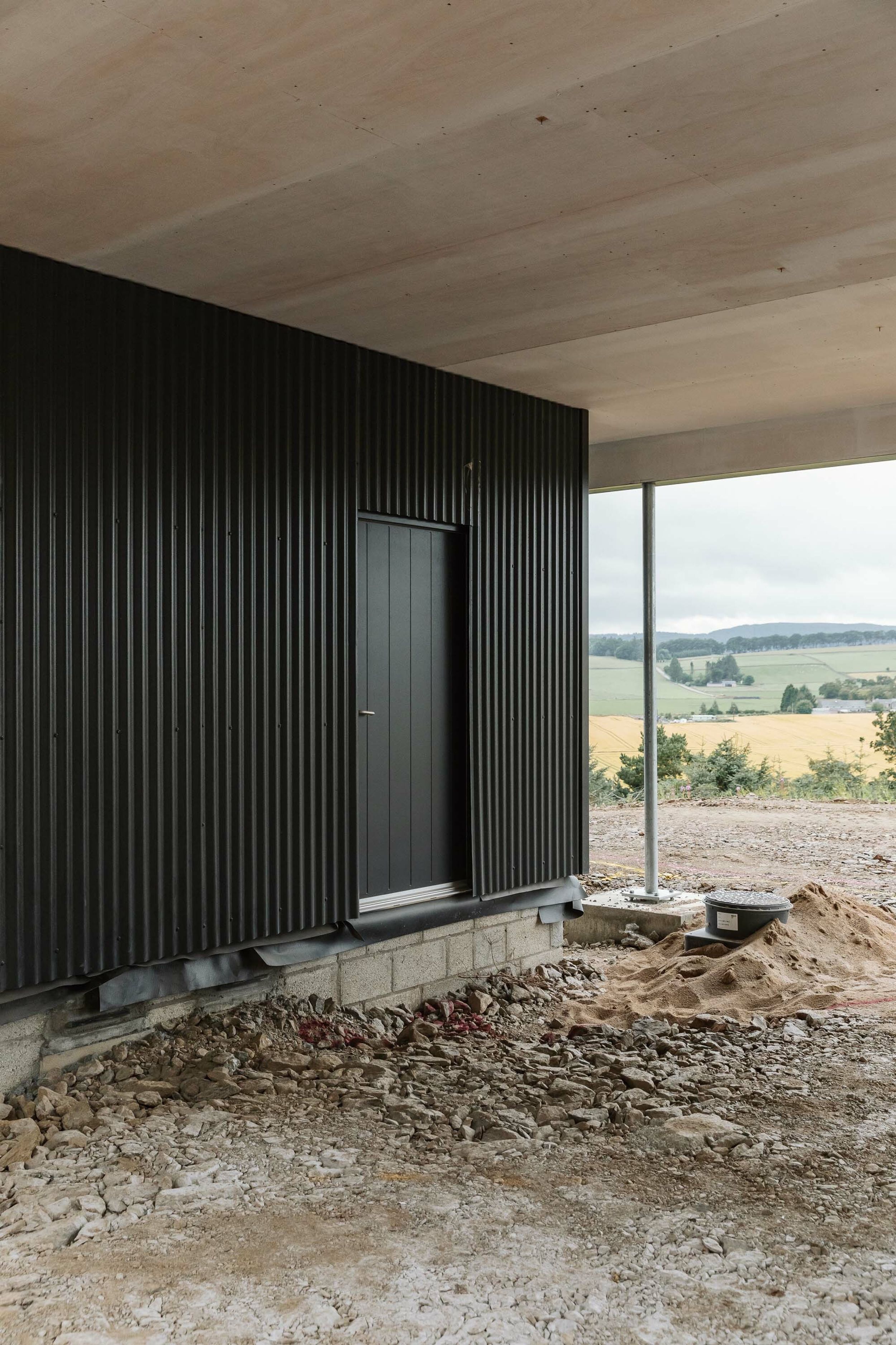 A partially-built home clad in grey corrugated sheeting. A black door is in the middle of the sheeting. Beyond the home, there is rural views of farmer’s fields. Rubble and builder’s sand can be seen in the foreground.