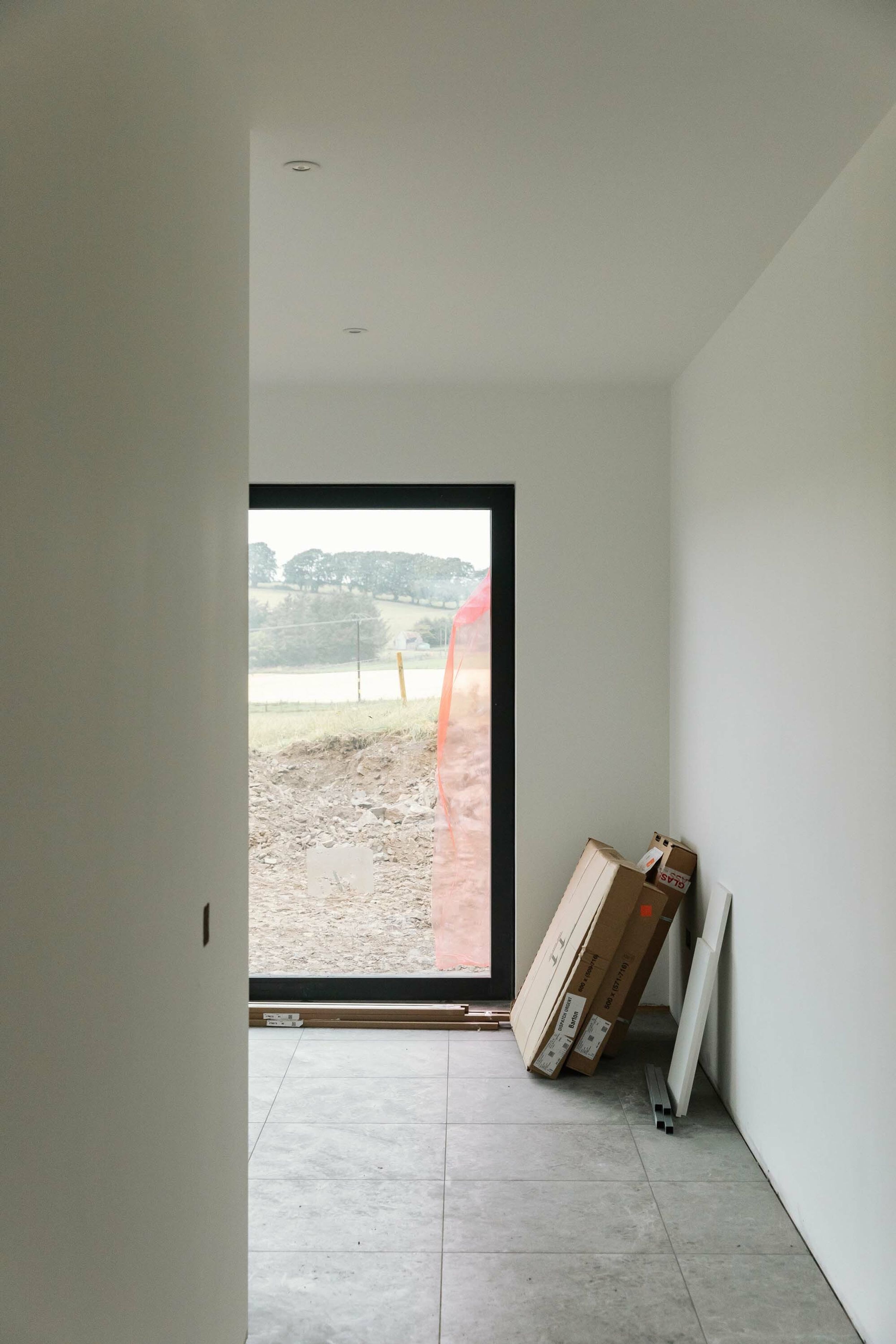 White freshly painted walls inside a partially built home. Grey tiles. A black sliding doors is at the end of a corridor. It reveals rural views.