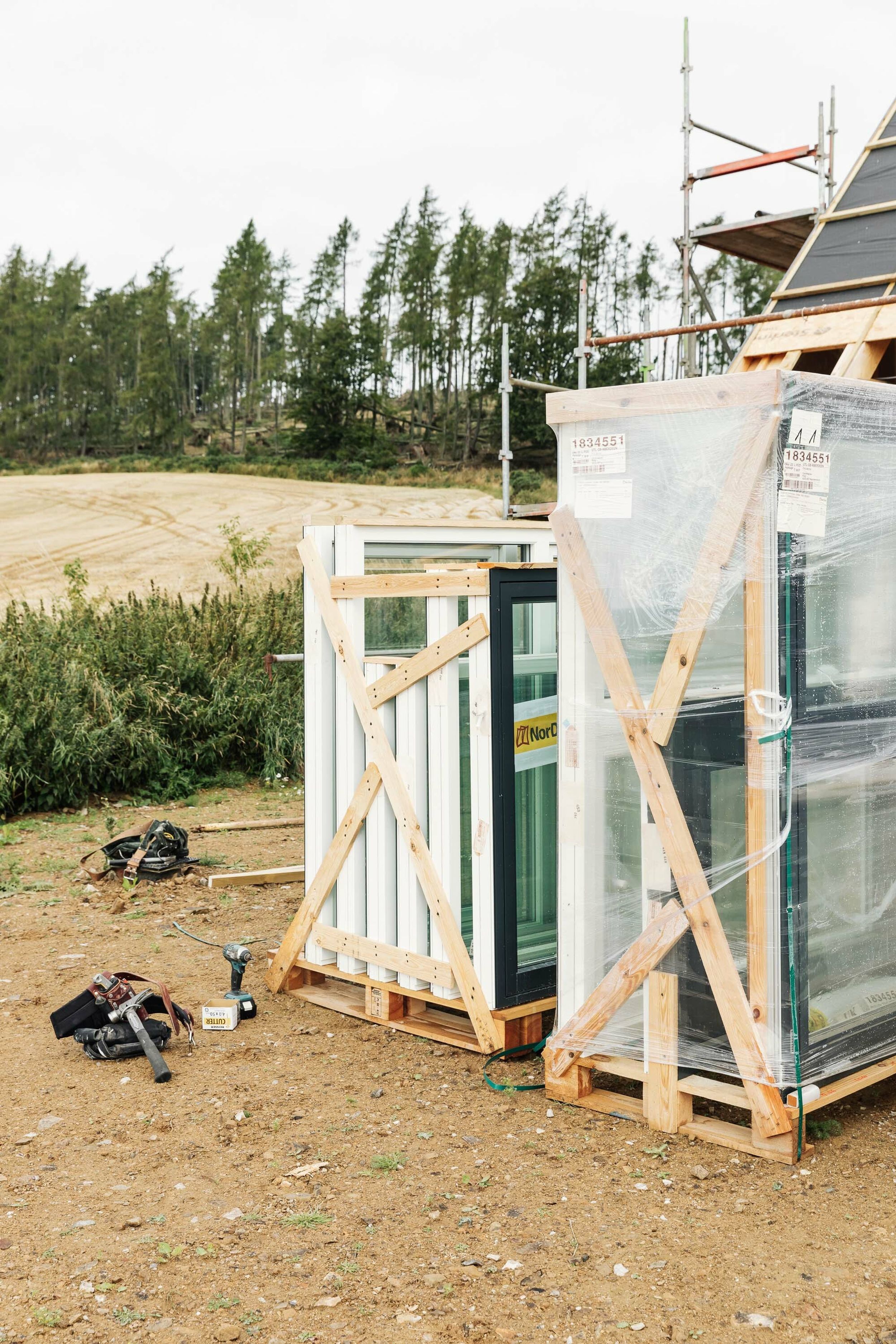 New Nordan windows are stacked on a crate, ready to be installed in a new build home. Forest and barley fields are in the background. The edge of a house and scaffolding can be seen.