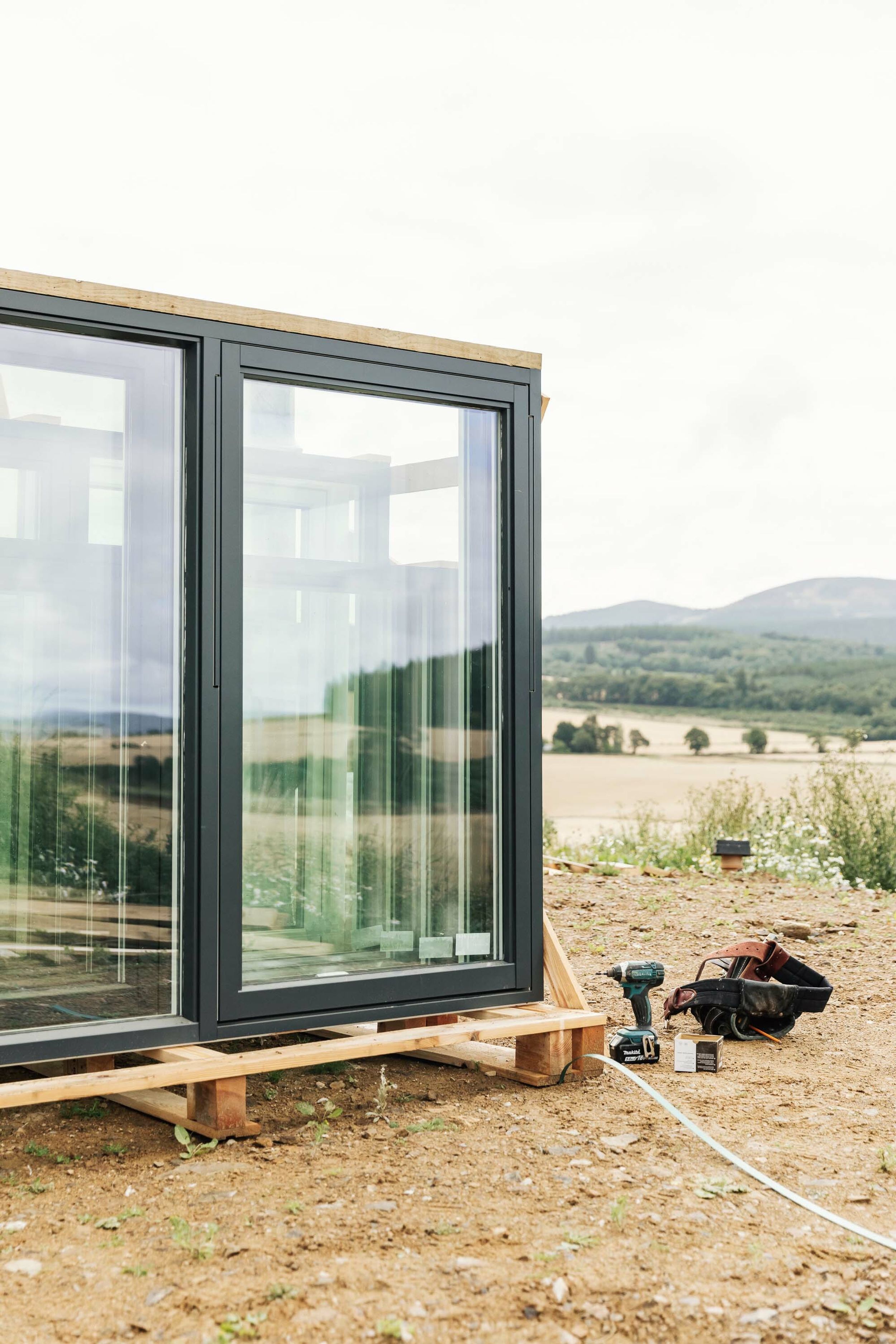 New Nordan windows are stacked on a crate, ready to be installed in a new build home. A drill and toolbelt lay on the bare earth beside the windows. In the distance, are barley fields, forest and hills.
