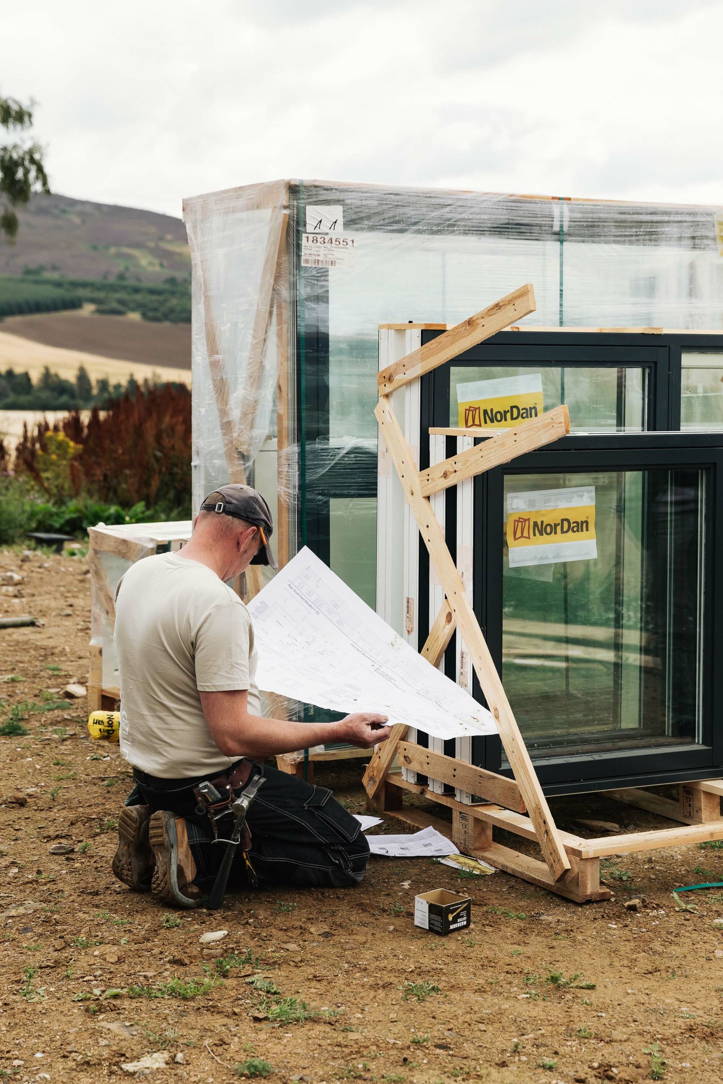 A builder kneels on the ground, next to a group of windows which are about to be installed. He holds and reads big, white plans.