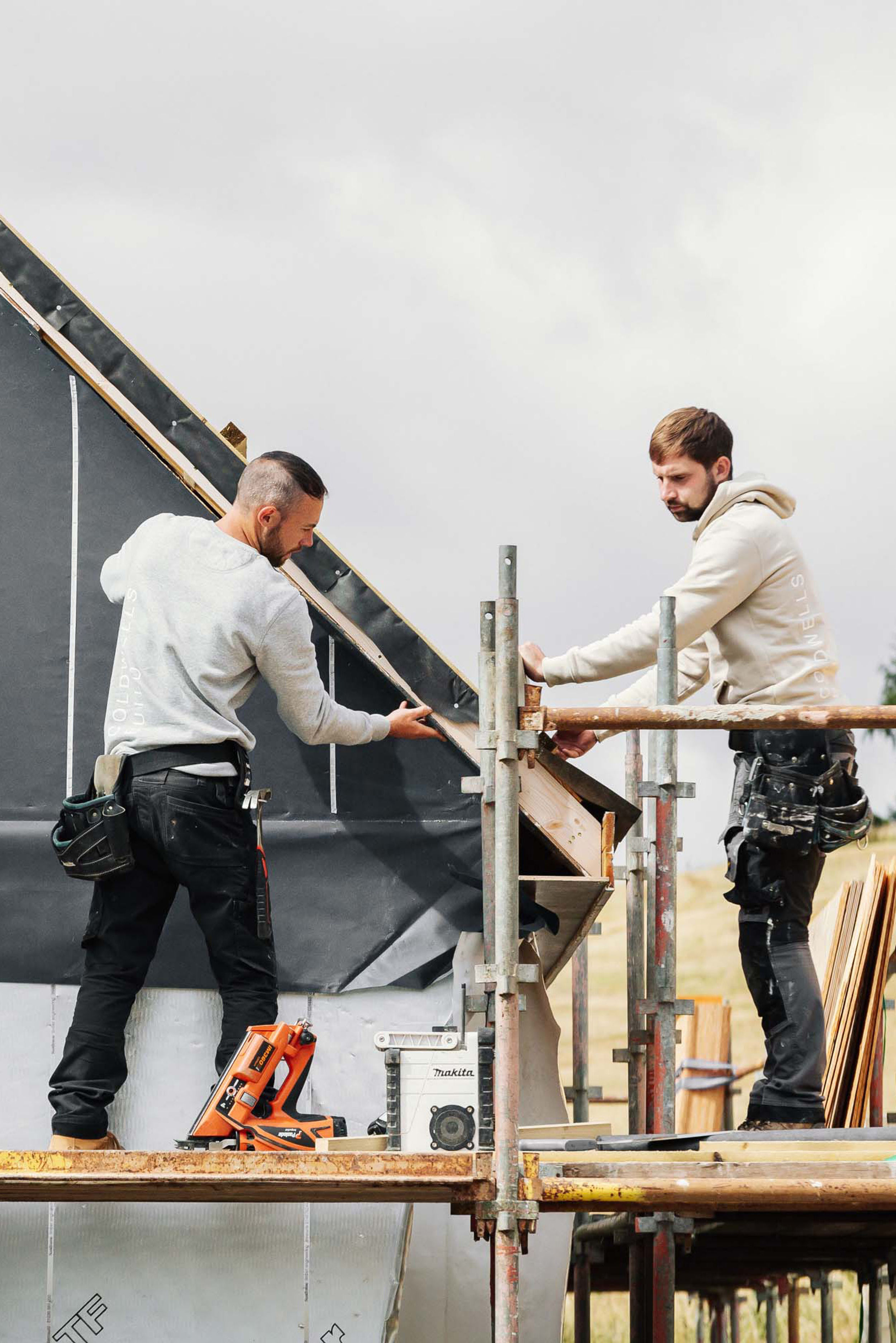 Two builders stand on scaffold and install black roofing felt on a new build home.