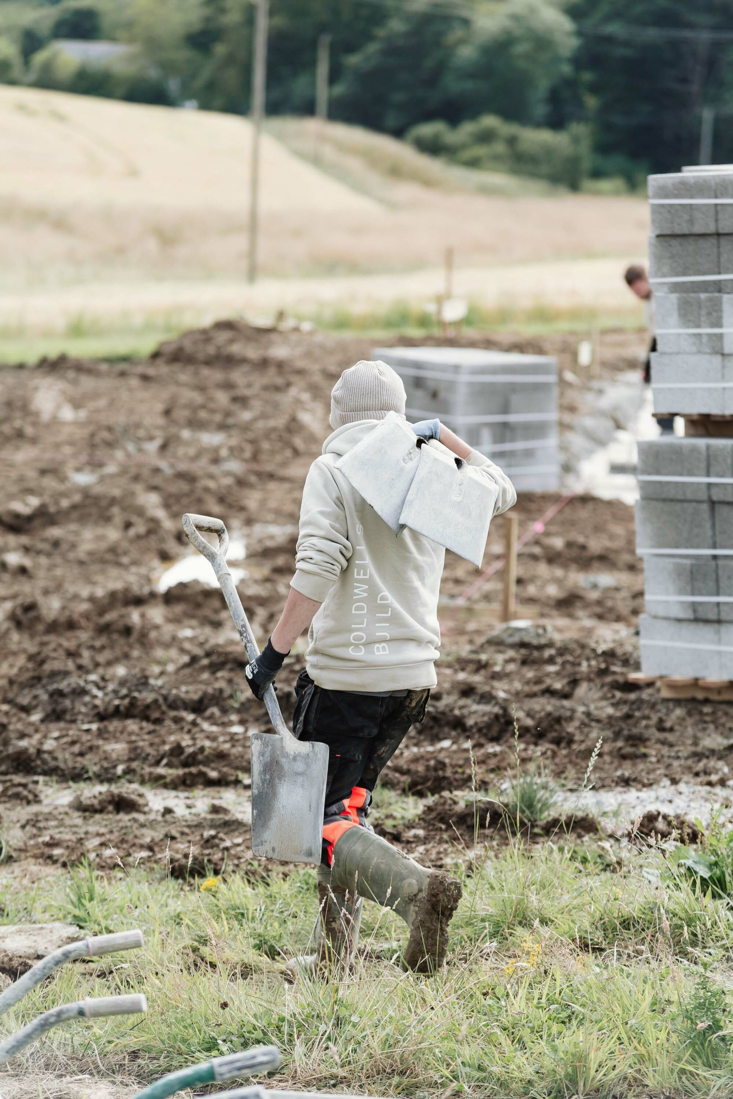 A builder wearing muddy boots carries two spades across his shoulder and grasps another in his hand.