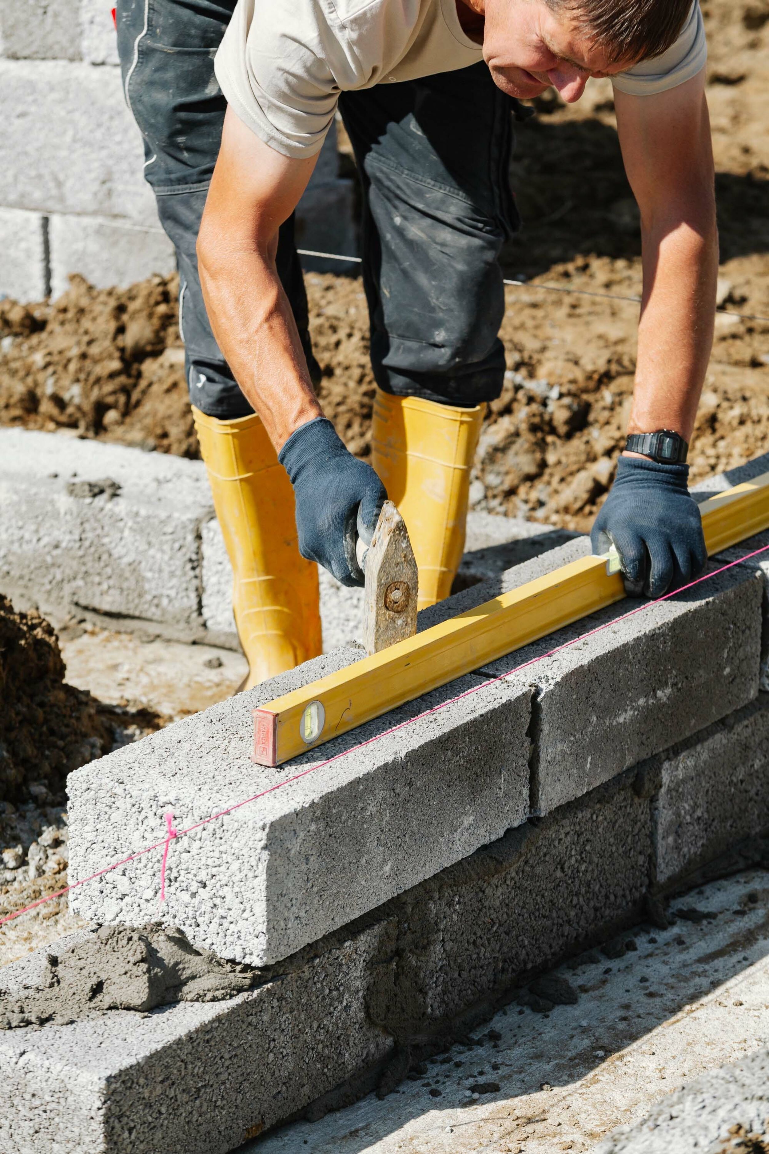 A construction worker holds a level against concrete blocks, freshly laid in wet cement, to build the finished floor height of a new build home. He uses a tool to chap the blocks into place and get them level.