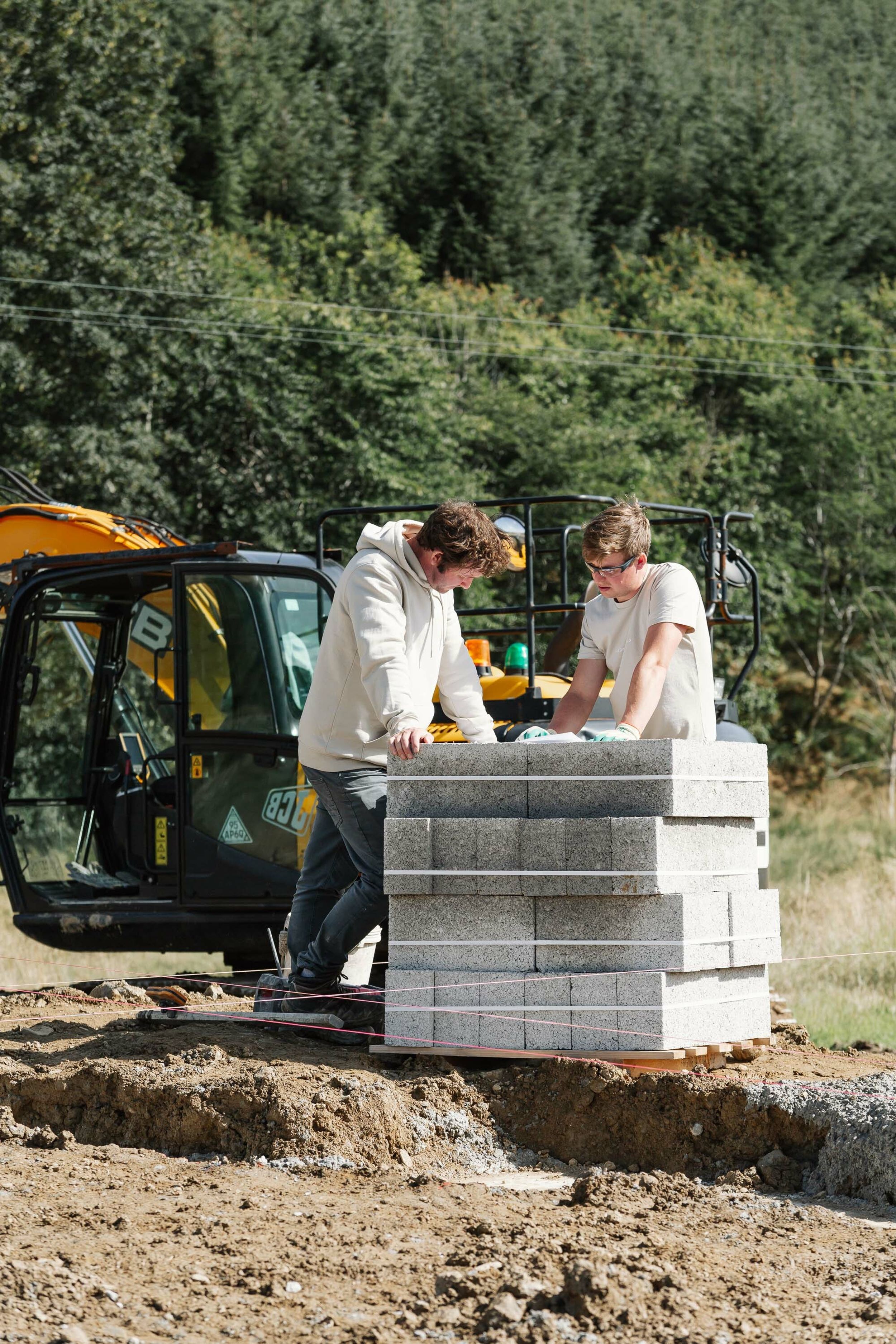 Two builders stand at the site of a new build home. They read large, white plans which are resting on a pile of concrete blocks. A yellow digger is parked in the background.