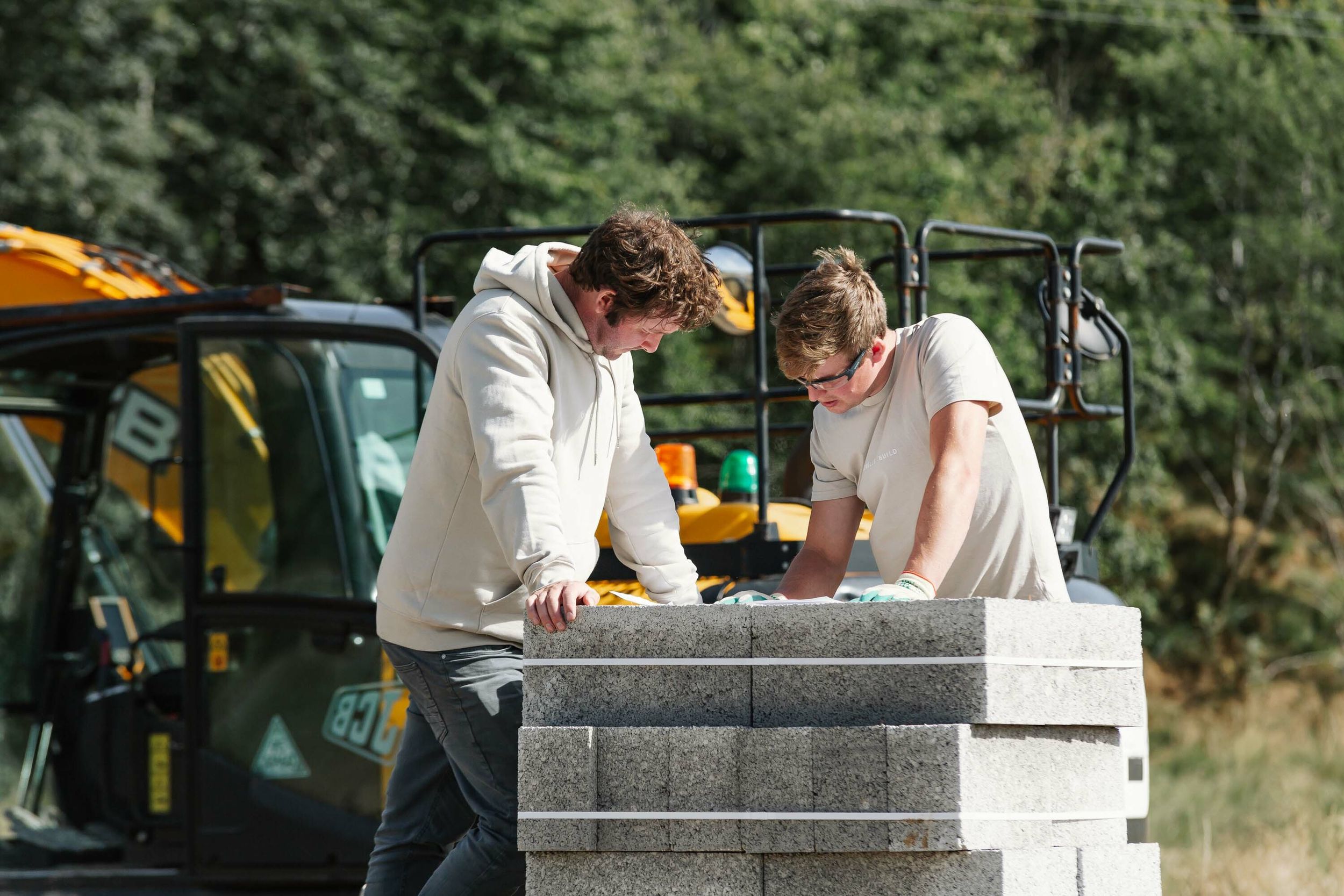 Two builders stand at the site of a new build home. They read large, white plans which are resting on a pile of concrete blocks. A yellow digger is parked in the background.