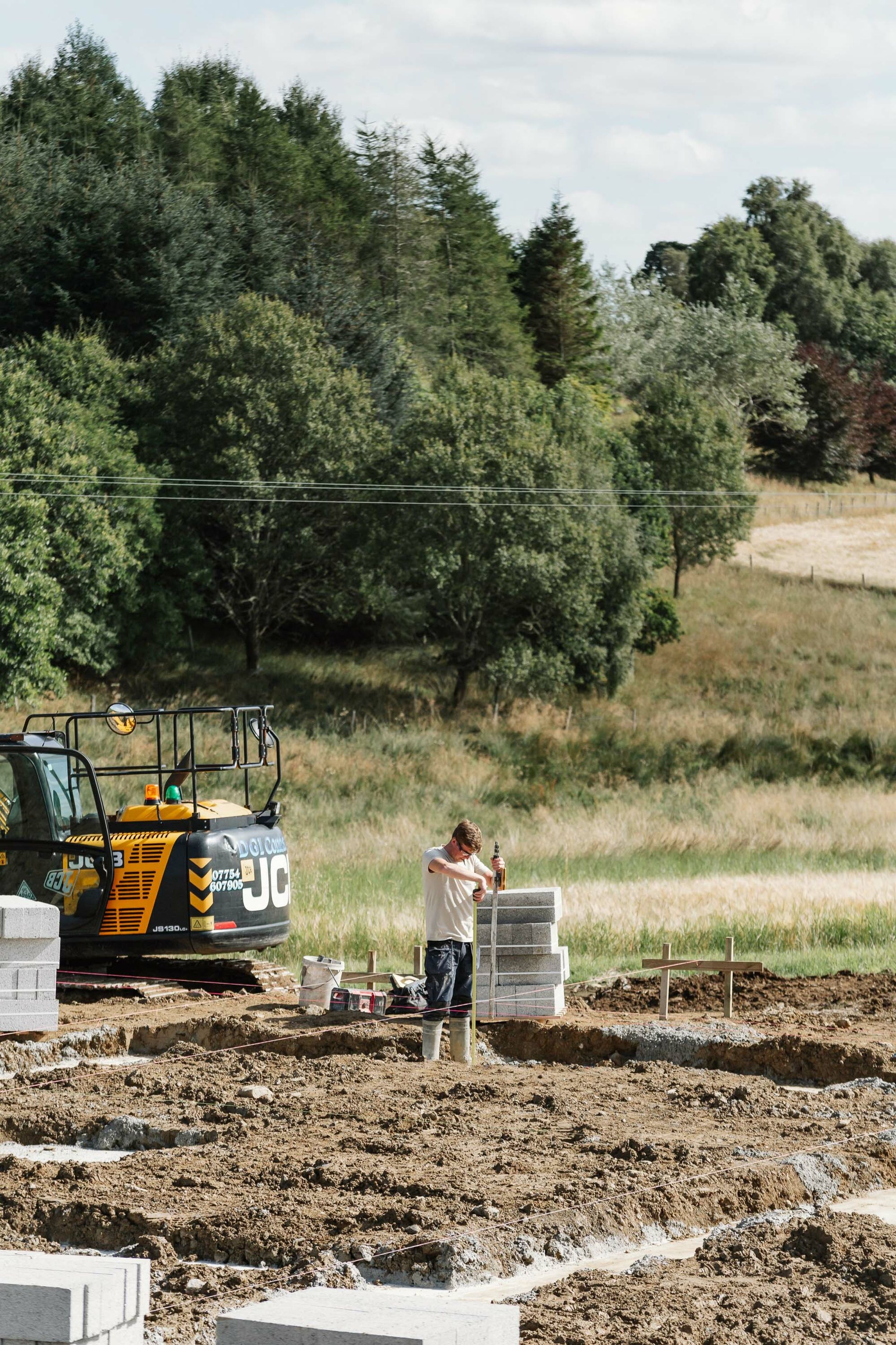 A builder measures out the foundations of a rural new build home. Trees and yellow digger are in the background.