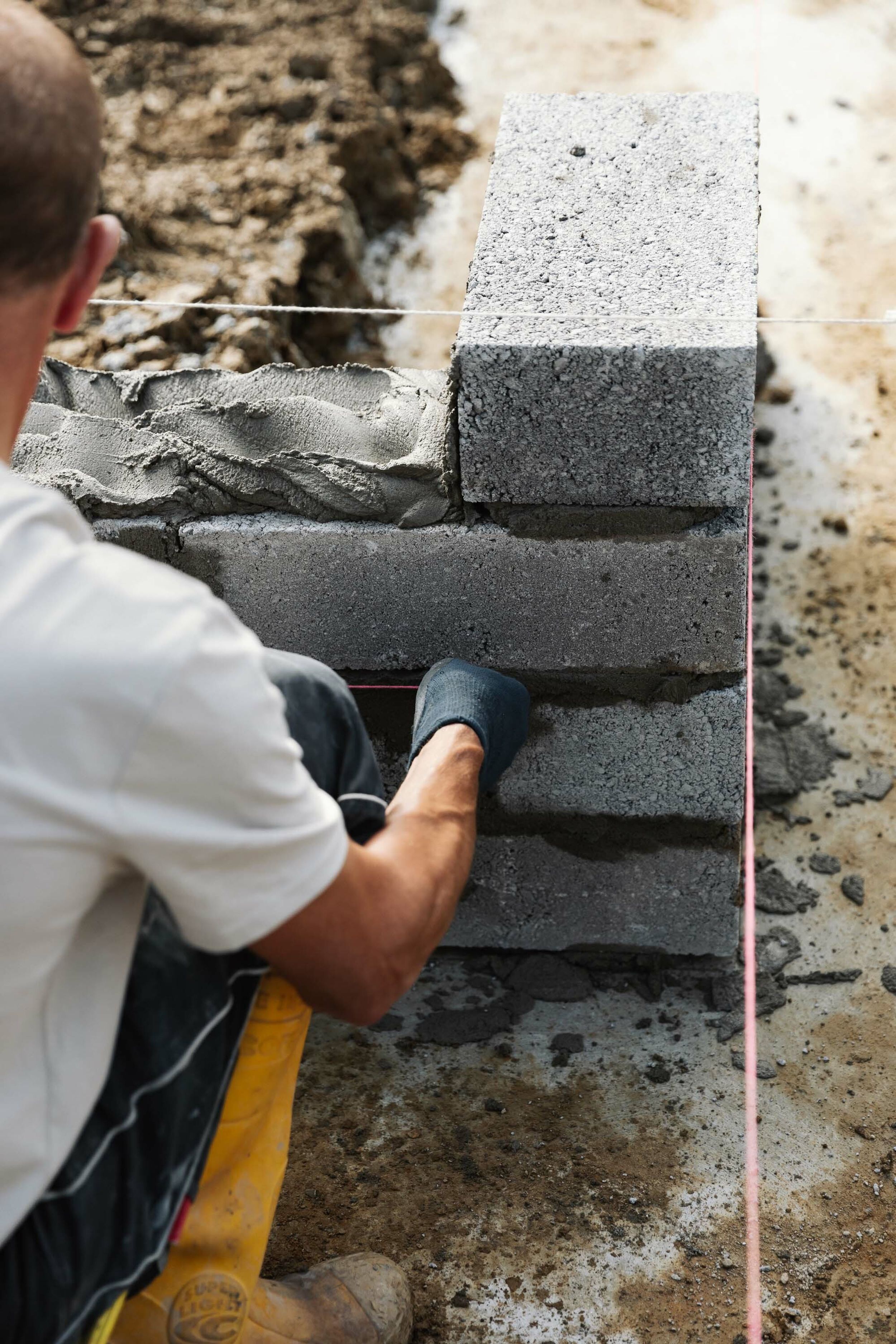 A builder crouches down to lay more blockwork to build the floor level of a new build home. He hold bright pink builders string in his hands. We cement has been dolloped on the blocks in front of him.