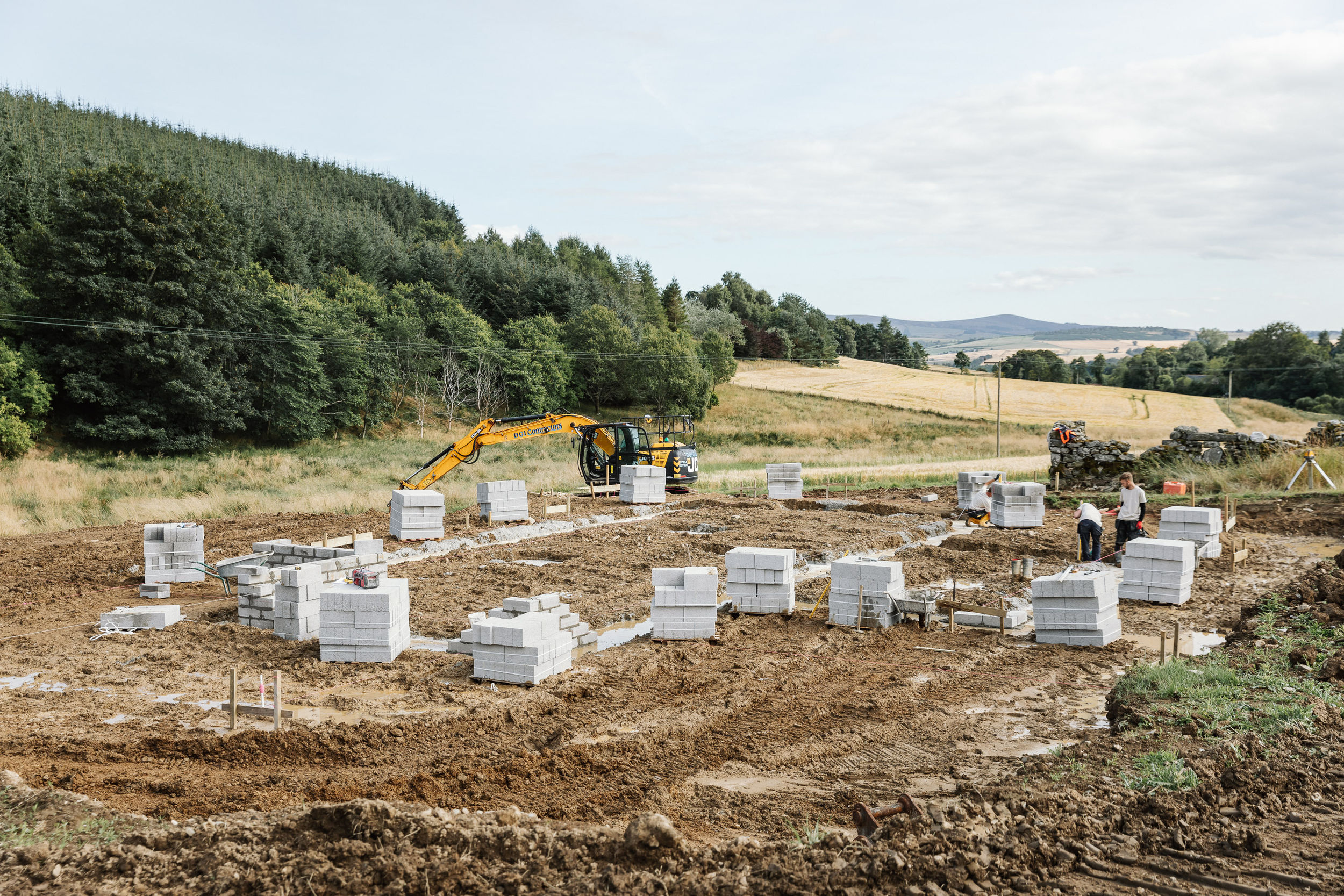 A yellow digger works on a construction site where a new house is being built. Concrete blocks are stacked in piles around the site. Two builders are speaking to each other. Forest surrounds the site.