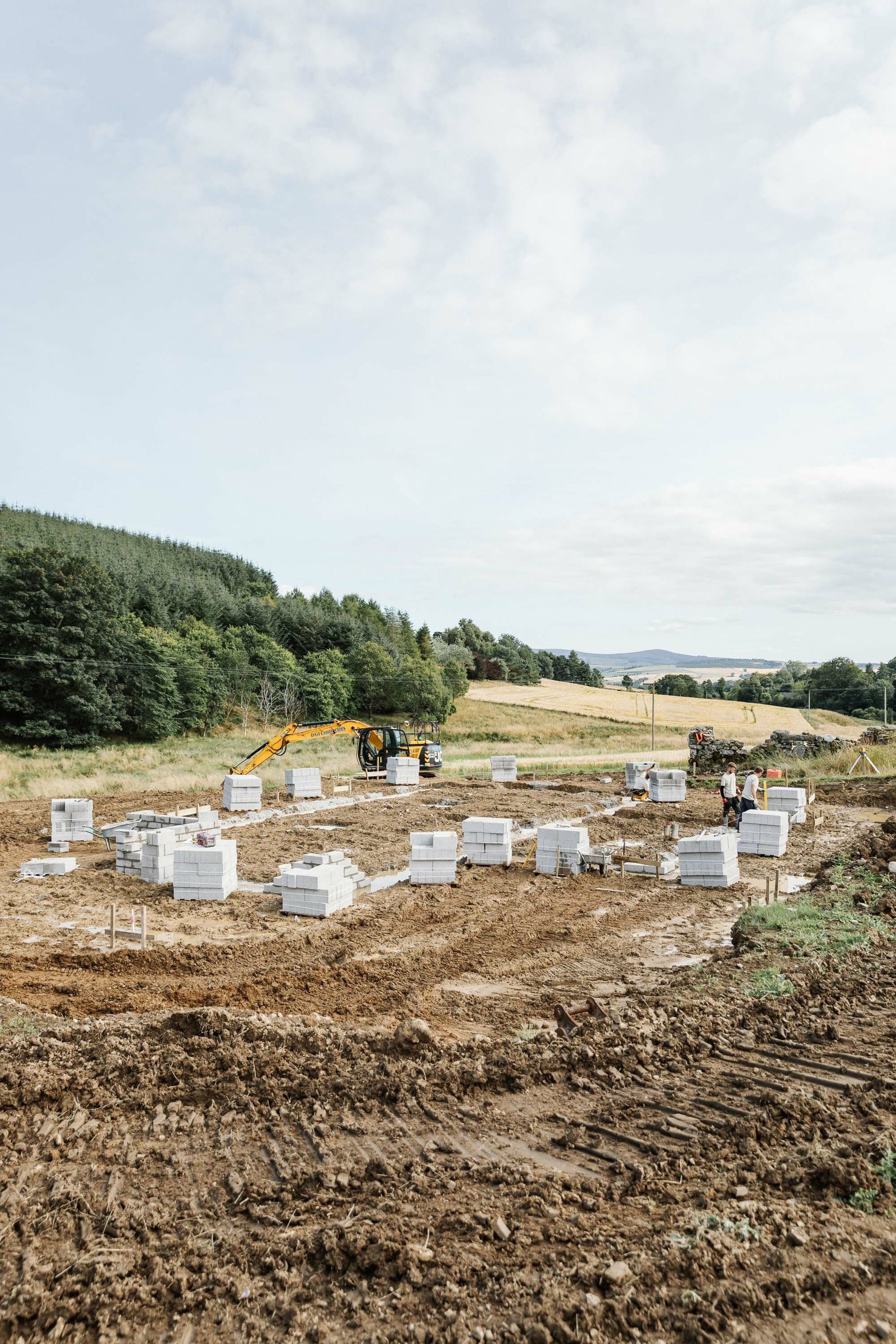 High angle shot of a muddy building site. The foundations of a new house are in place. Concrete builder’s blocks are stacked in pallets and surround the foundations, ready to build walls. A yellow digger, barley fields and forest is in the background.