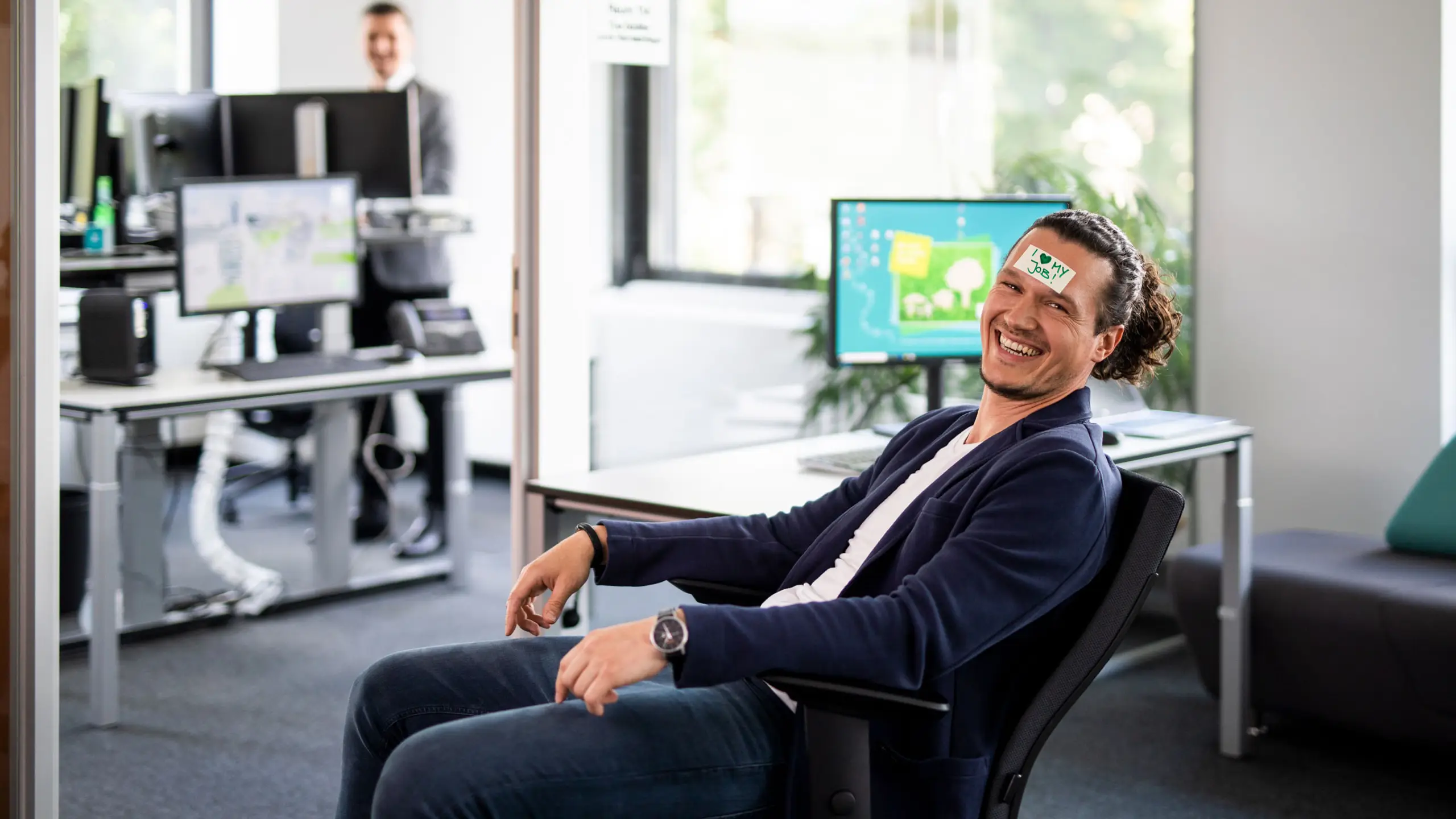 Mitarbeiter, der auf einem Bürostuhl sitzend in die Kamera lacht und ein Post-It mit "I love my job" auf der Stirn kleben hat