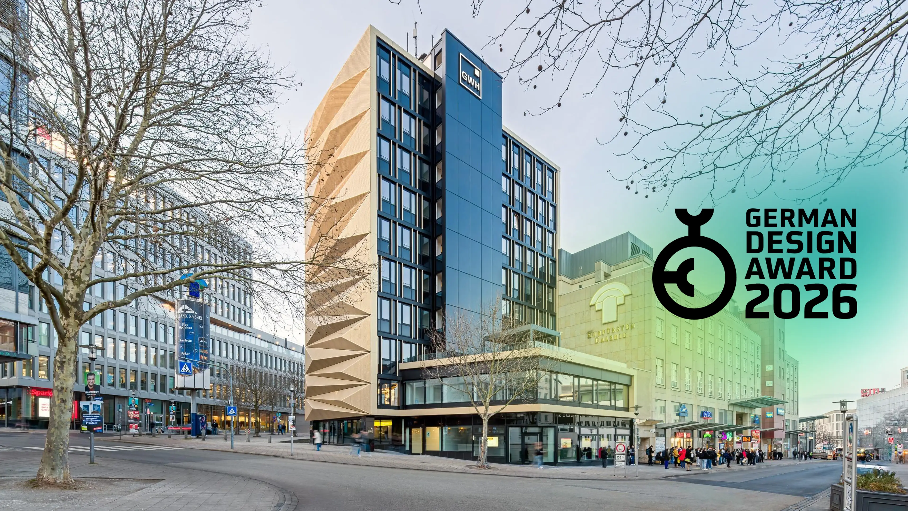 Modernes mehrstöckiges Bürogebäude an einer Straßenecke mit Bäumen und Passanten im urbanen Umfeld, Logo des German Design Award 2026 rechts im Bild