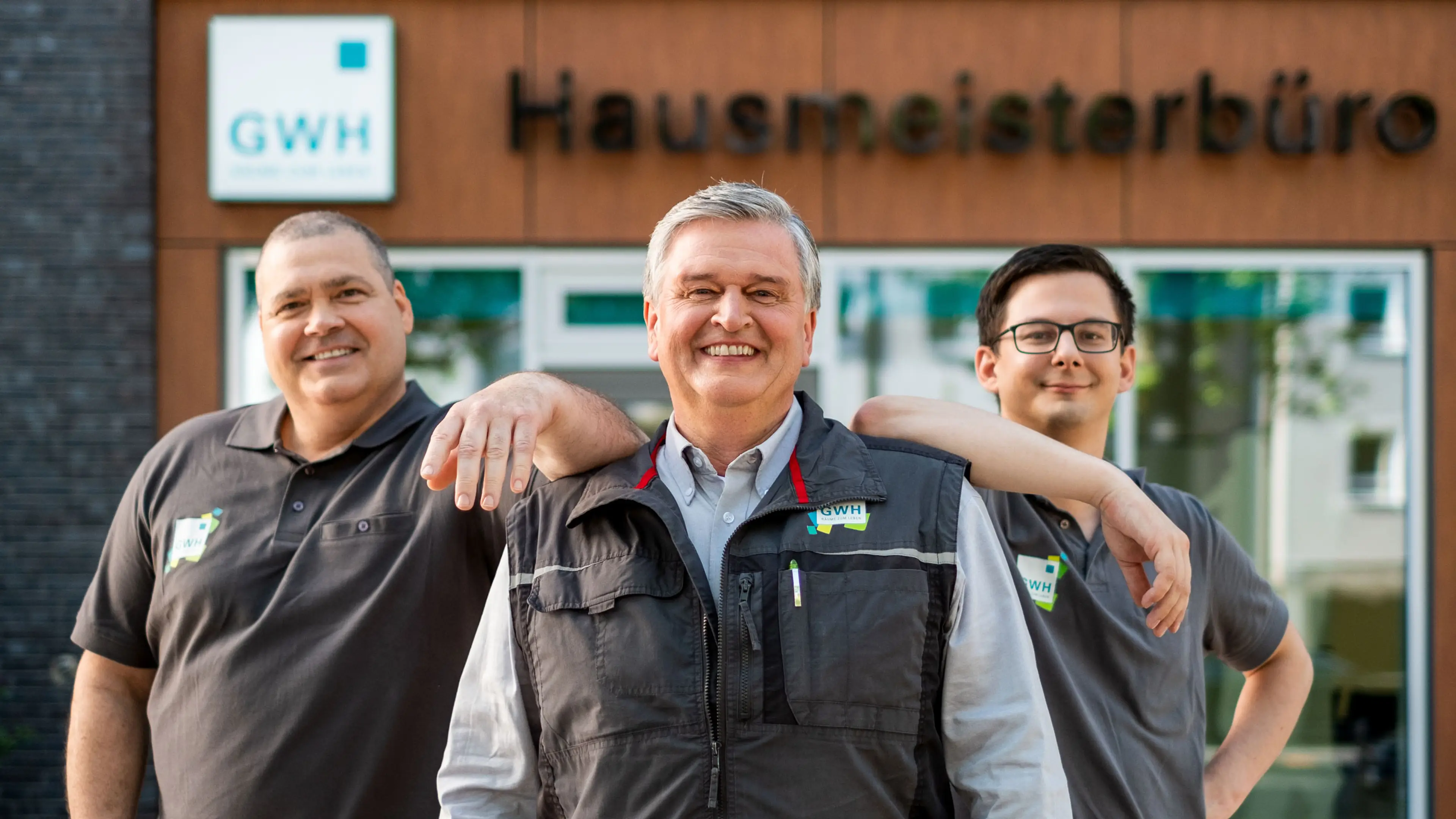 Drei Hausmeister stehen vor dem Hausmeisterbüro und lachen in die Kamera
