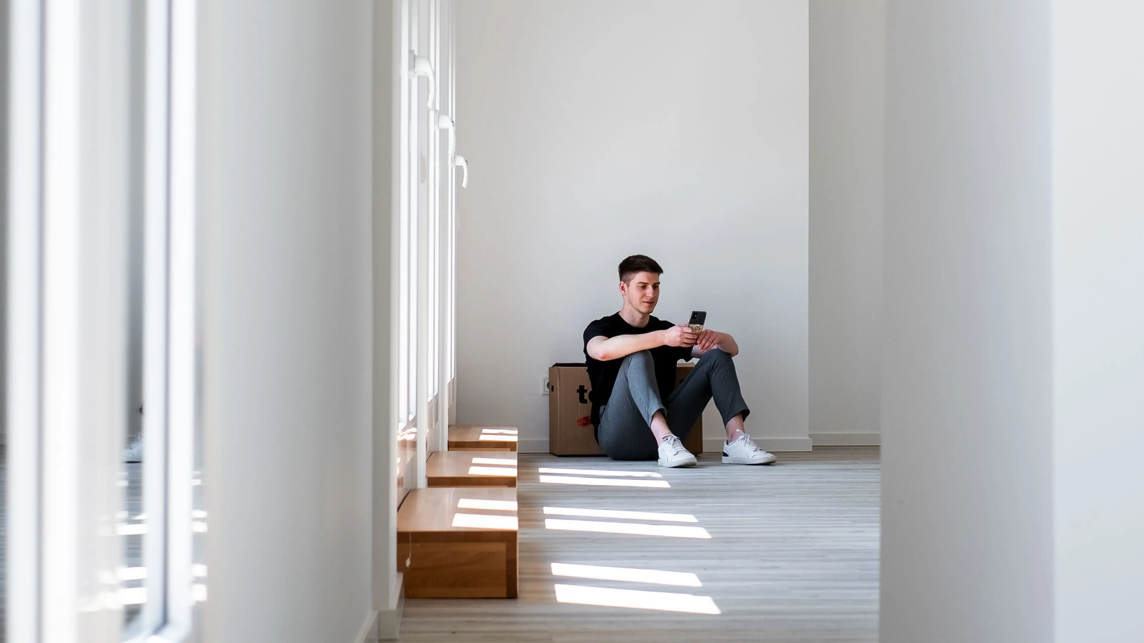 Ein Mann sitzt an einen Umzugskarton gelehnt in seiner neuen Wohnung und schaut auf sein Handy