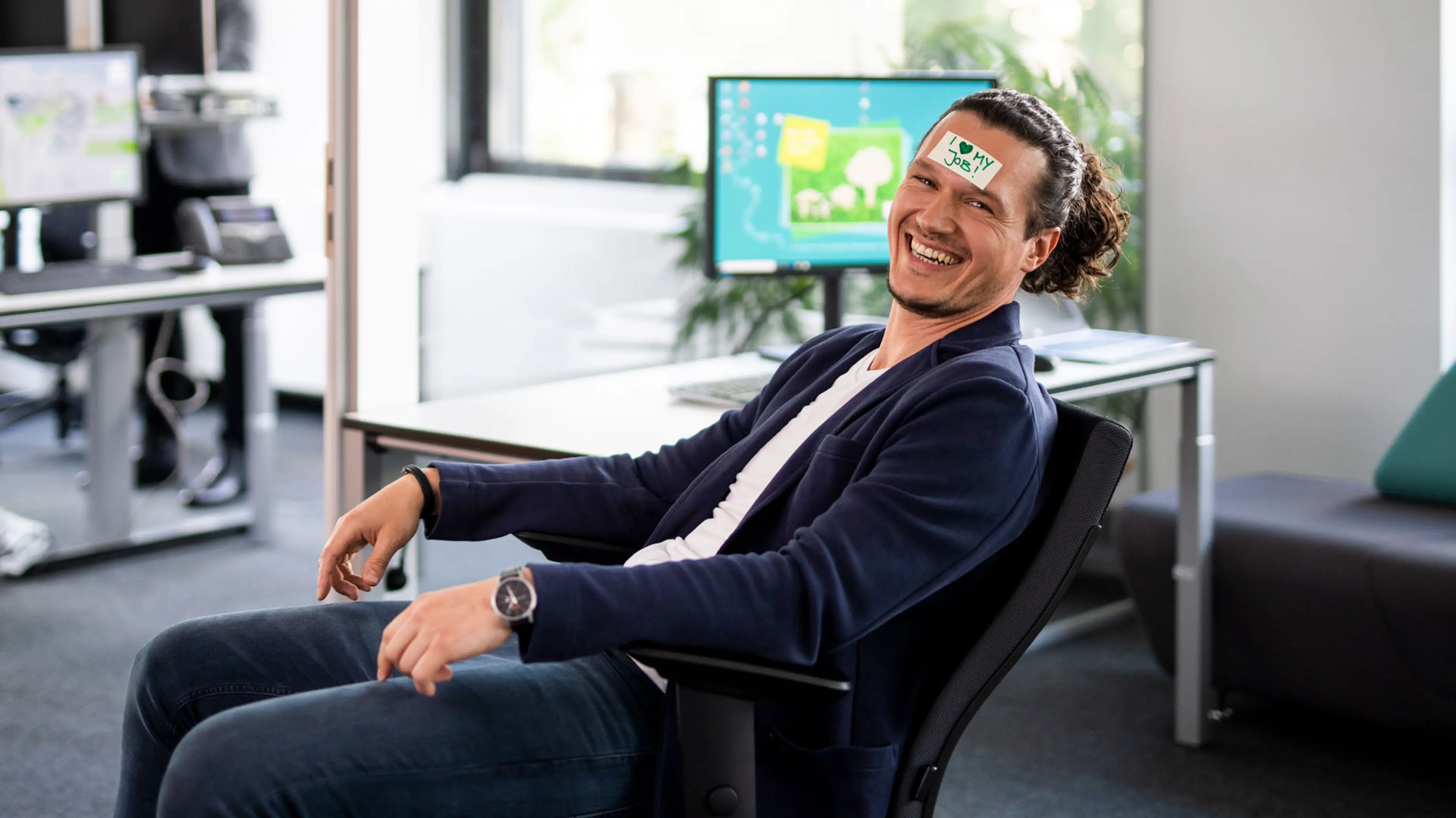 Mitarbeiter, der auf einem Bürostuhl sitzend in die Kamera lacht und ein Post-It mit "I love my job" auf der Stirn kleben hat