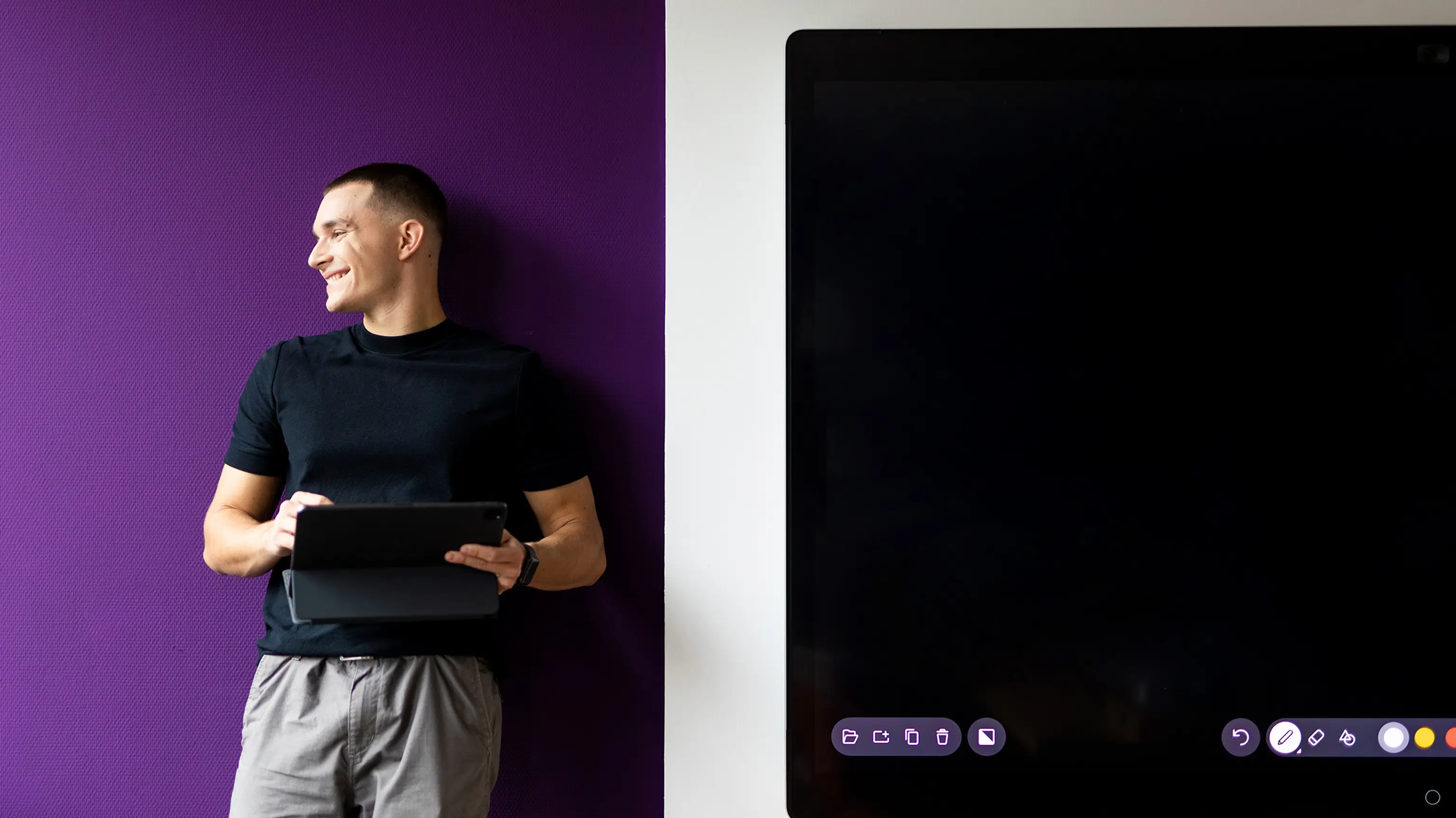 Auszubildender lehnt mit iPad in der Hand an der Wand eines Besprechungsraums neben einem digitalen Board