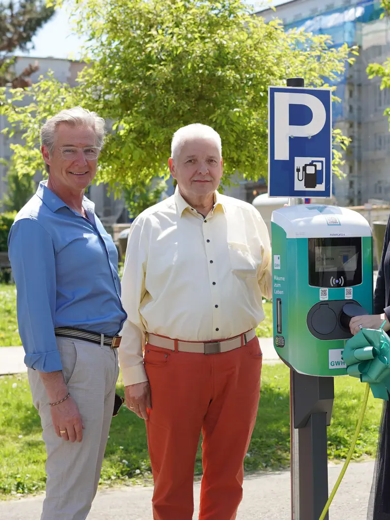 Drei Personen stehen an einer Ladesäule für Elektrofahrzeuge, eine Person hält das Ladekabel an ein geparktes Auto