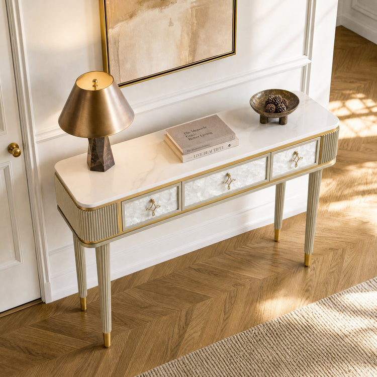 A three-drawer console has a white veined top, ribbed legs, patterned fronts, and warm-metal trim