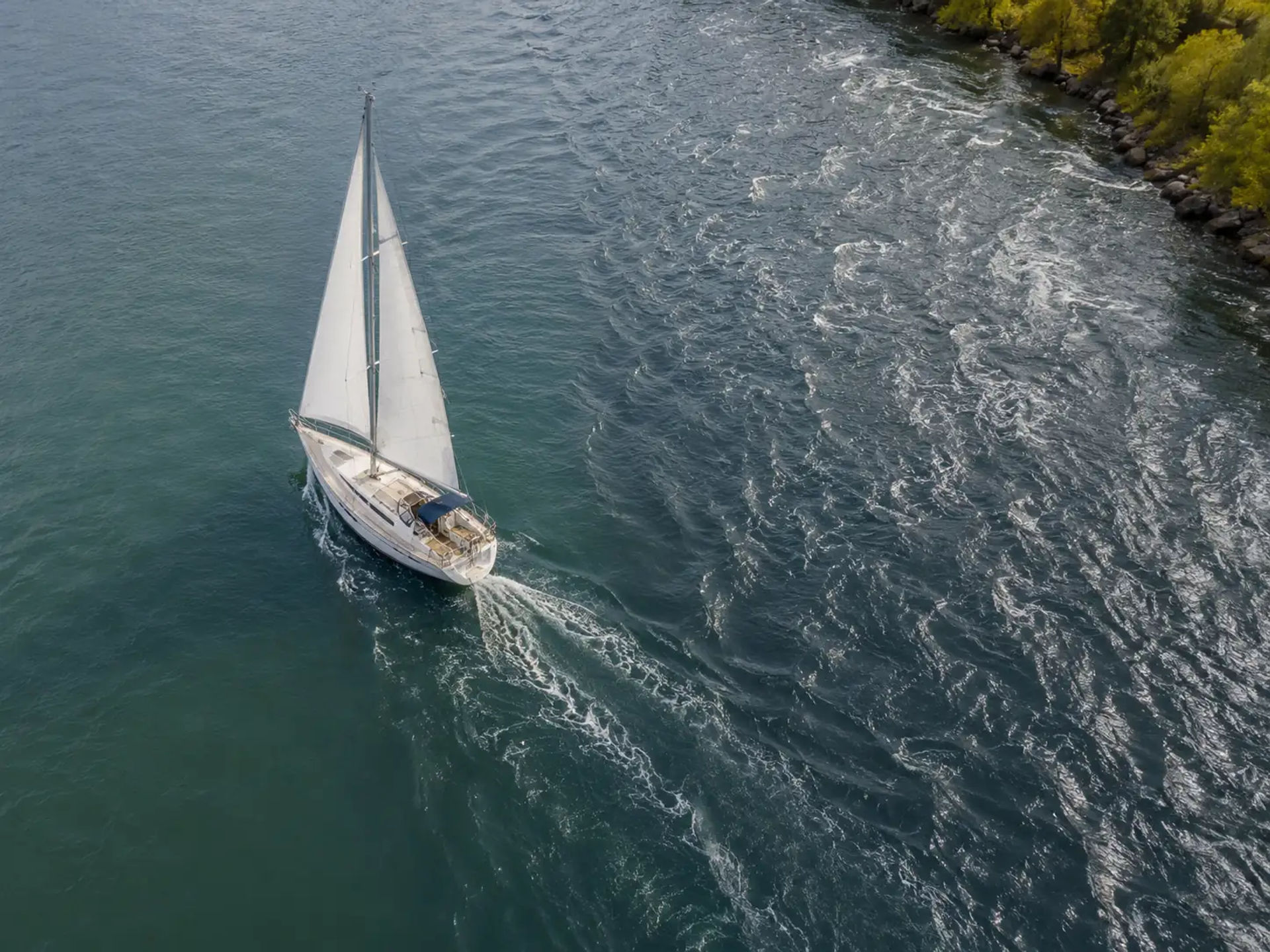 Sailboat crossing from calm water into turbulent, choppy water near a rocky shoreline