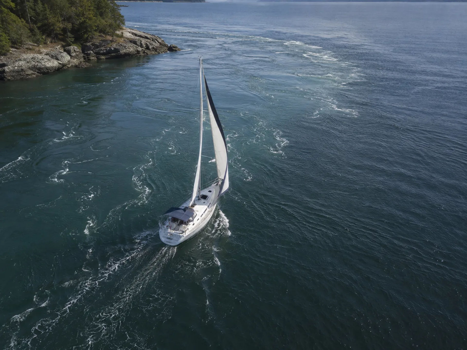 Sailboat sailing through turbulent tidal current near a rocky, forested coastline