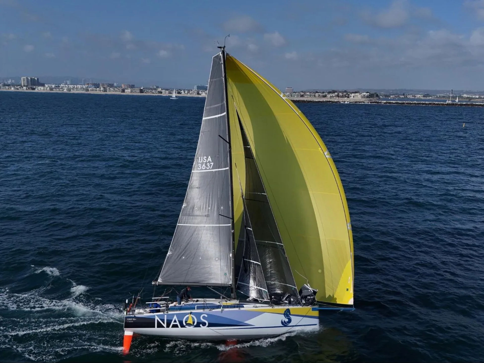 Naos racing sailboat sailing with a large yellow spinnaker near the coast