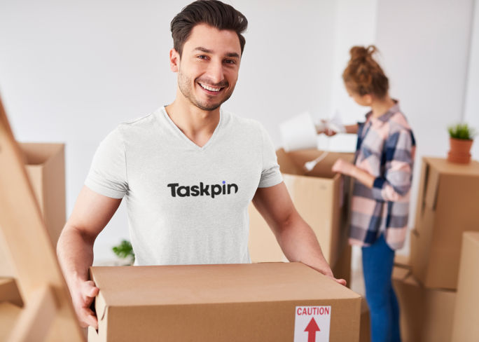 A smiling man in a white t-shirt with the words Taskpin on it is moving a cardboard box for moving