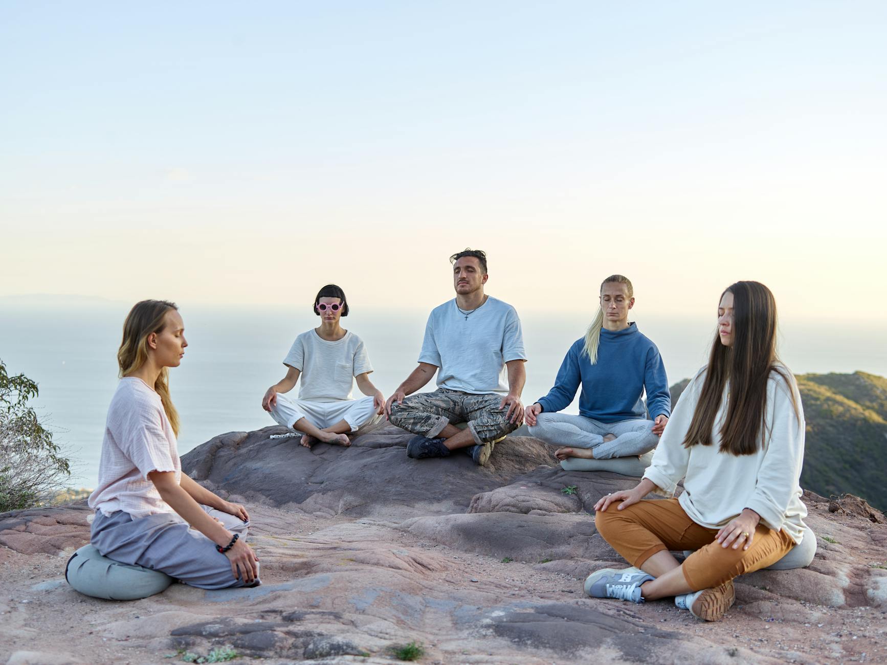 Smiling small wellness group meditating outdoors beside solo traveler journaling under tree