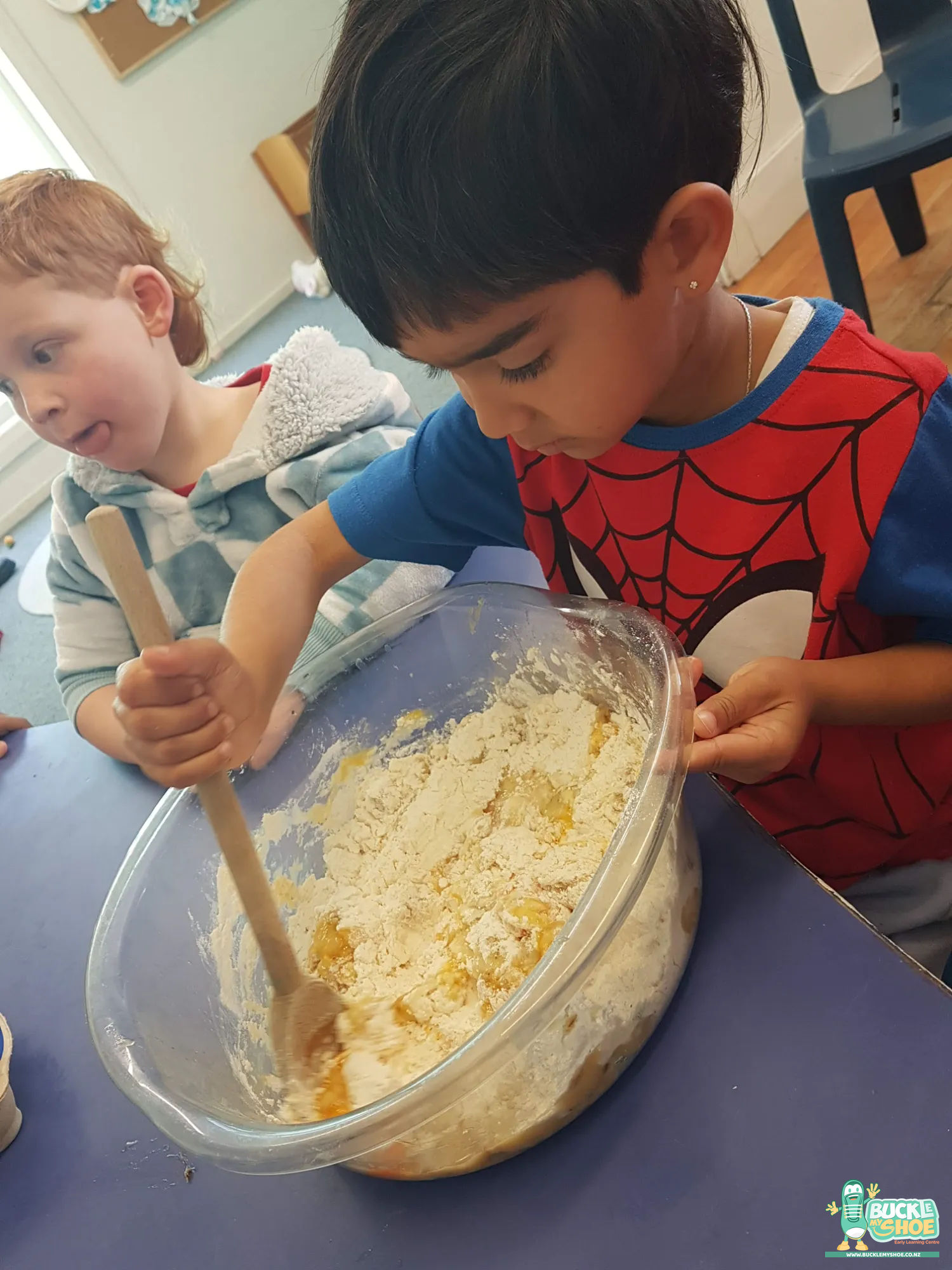 Buckle My Shoe Tauranga & Papamoa | Childcare, Preschool, Daycare | A teacher helping a child pour milk into a large mixing bowl.