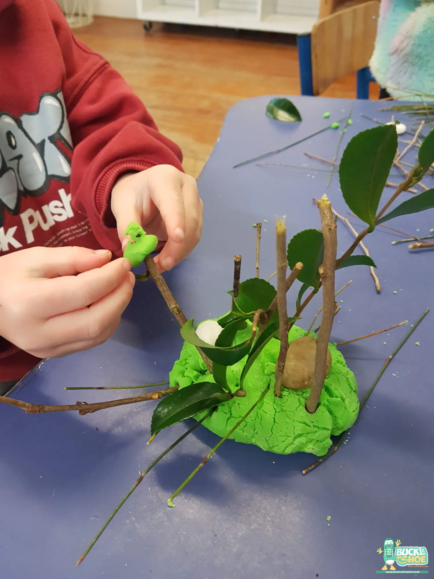 Buckle My Shoe Tauranga & Papamoa | Childcare, Preschool, Daycare | A child presses a twig into playdough to create a tree.