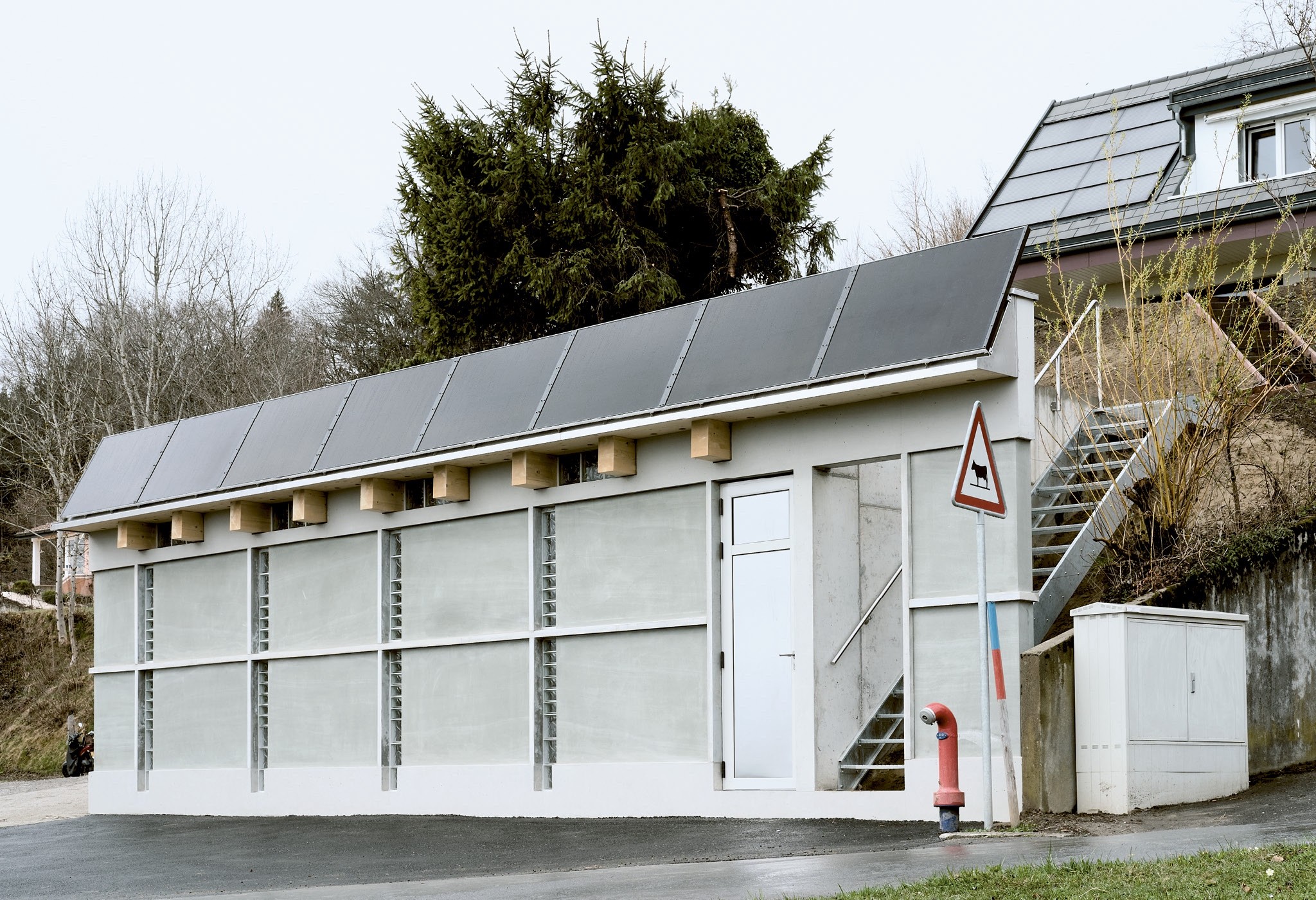 Die Fassade der Garagenerweiterung besteht aus in Beton gefassten Quadern aus lokalem Molasse-Stein und trägt ein Gesims aus Standard-Solarmodulen.