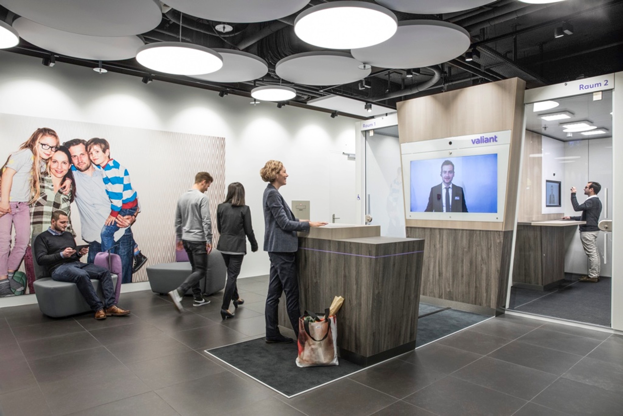 Am digitalen Empfang begrüsst ein Mitarbeiter der Bank die Kundin, im hinteren Bereich des Raums schliessen die Self-Banking-Räume und die Beratungszimmer an.
