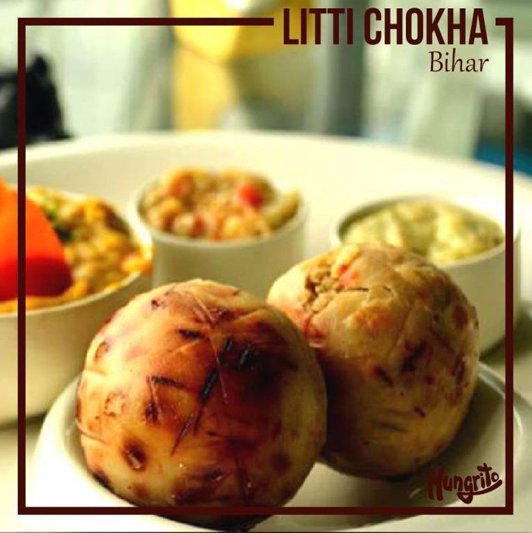 Two round, golden brown Litti balls with a bowl of spicy Chokha in the background.