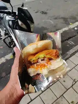 Vada Pav You’ll Regret Not Trying - Best Vada Pav in Mumbai 5 A delectable Vada Pav from Bhau Vada pav in tin foil. In the background there is a bike, road and footpath.