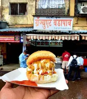 Vada Pav You’ll Regret Not Trying - Best Vada Pav in Mumbai 7 An exquisite Vada Pav served in paper dish held in hand while the background has the sign of Graduate Vada Pav and gives the crowded glimpse of the stall.