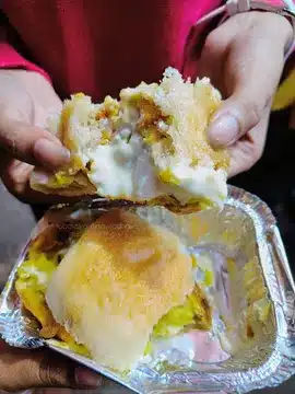 Vada Pav You’ll Regret Not Trying - Best Vada Pav in Mumbai 10 Delicious Khidki Vada' Vada Pav served in a container. There are two Vada Pavs in frame; one is laying in container and the other is being pulled apart by two hands releasing or showcasing the cheese between them.