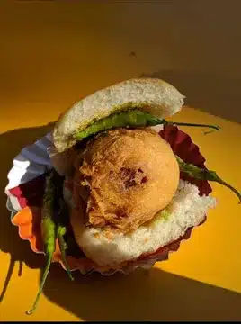 Vada Pav You’ll Regret Not Trying - Best Vada Pav in Mumbai 3 Parleshwar Vada Pav Samrat's appealing Vada Pav in a yellow background with Soft side light. The Vada Pav is served in a typical plate with Green Chilli.