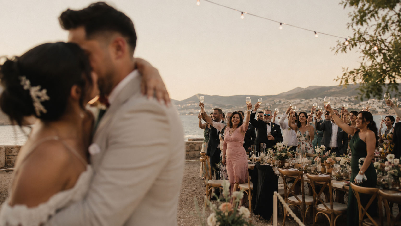 Foto documental de boda con pareja latina abrazándose fuera de foco mientras los invitados brindan alrededor, luz cálida de atardecer