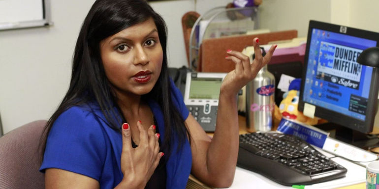 Kelly Kapoor with her hand on her chest at her desk in The Office