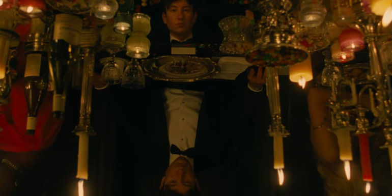 Oliver (Barry Keoghan) sitting at a grand table setting looking at his own reflection in Saltburn.