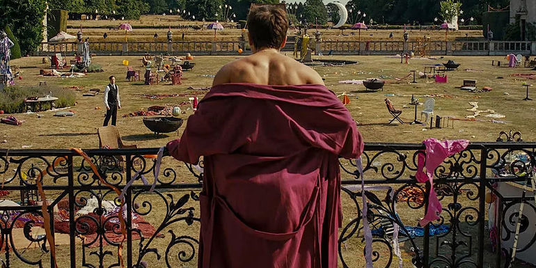 A shot of Oliver (Barry Keoghan) from behind, wearing a robe while looking out at the messy lawn on the Saltburn estate grounds in Saltburn.