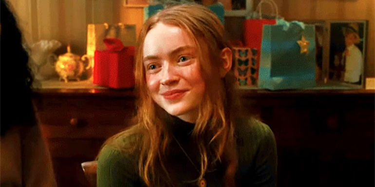 Sadie Sink as Her, sitting and smiling in Taylor Swift's All Too Well short film.