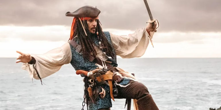 A cosplayer dresses as Captain Jack Sparrow, in outrageous garb with dredlocks and pirate hat, and gun belt laden with weapons, wielding a sword while tottering across rocks with the sea behind