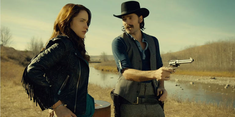 Doc Holliday holding a pistol next to Wynonna Earp on the side of a river bank