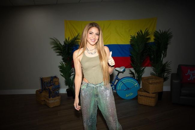 Shakira giving a thumbs-up in front of a Colombian flag, clad in a tank top and glittery pants, with indoor plants and a stationary bike in the background.