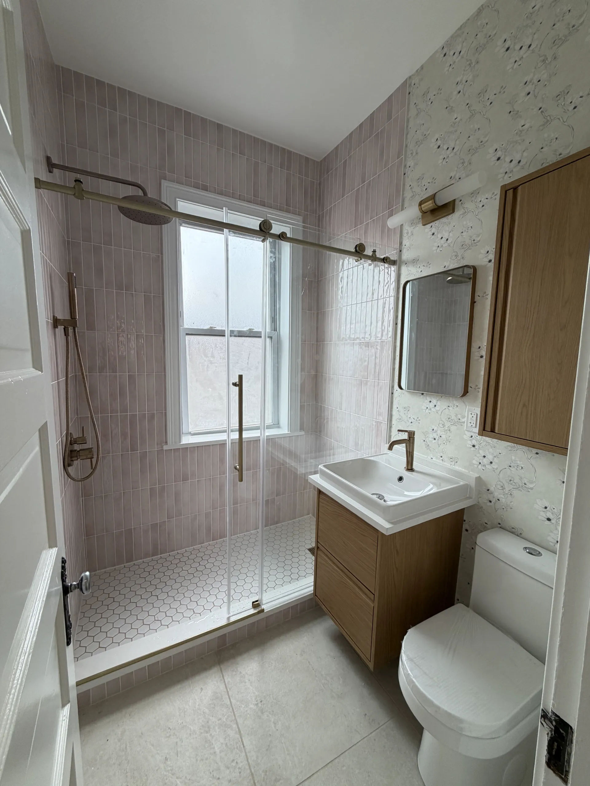 After photo of walk-in shower with pink zellige-style tile walls, white hex mosaic floor, frameless glass door, brass fixtures, and floating oak vanity in Chicago bathroom remodel by Assembly Squad